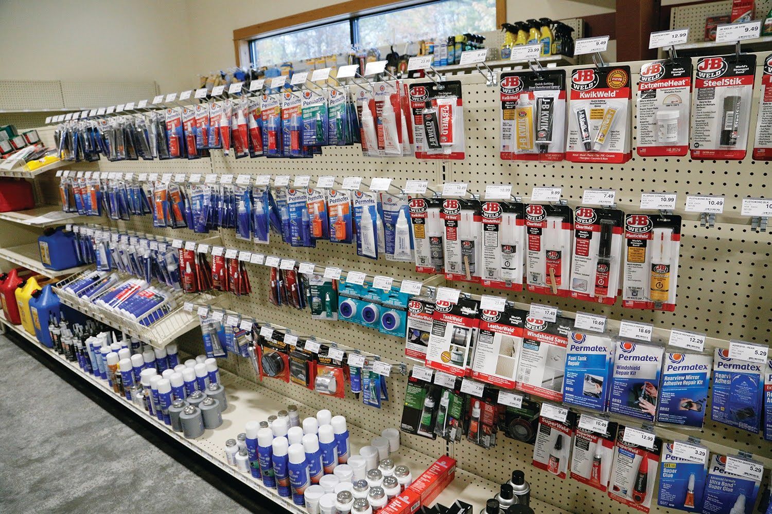 A retail aisle displays rows of small, packaged hardware supplies like adhesives and sealants hanging on a pegboard wall.