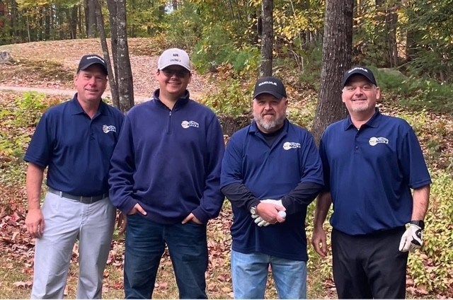 Four people in matching dark blue logo shirts and caps pose outdoors in a wooded area during autumn.