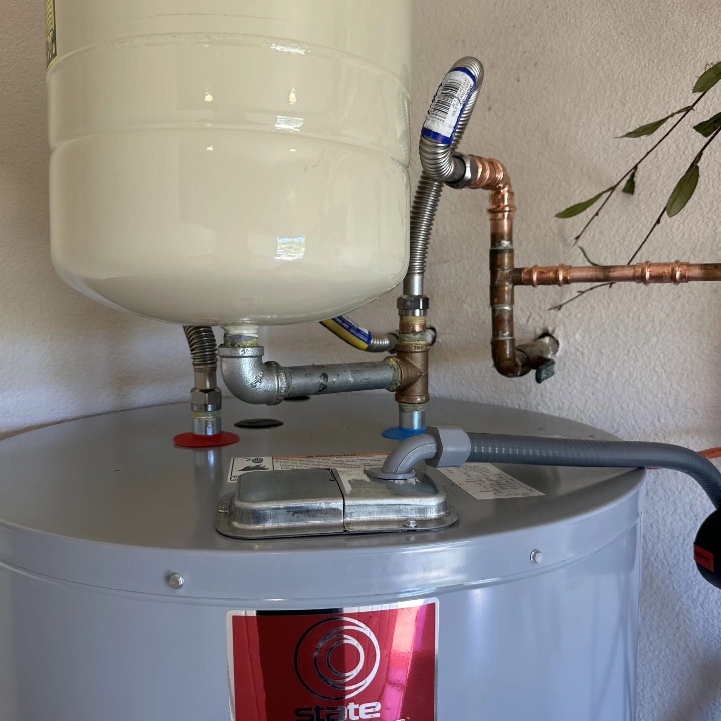 Plumbing and pipes on top of a white water heater with a red label and gray hose attached