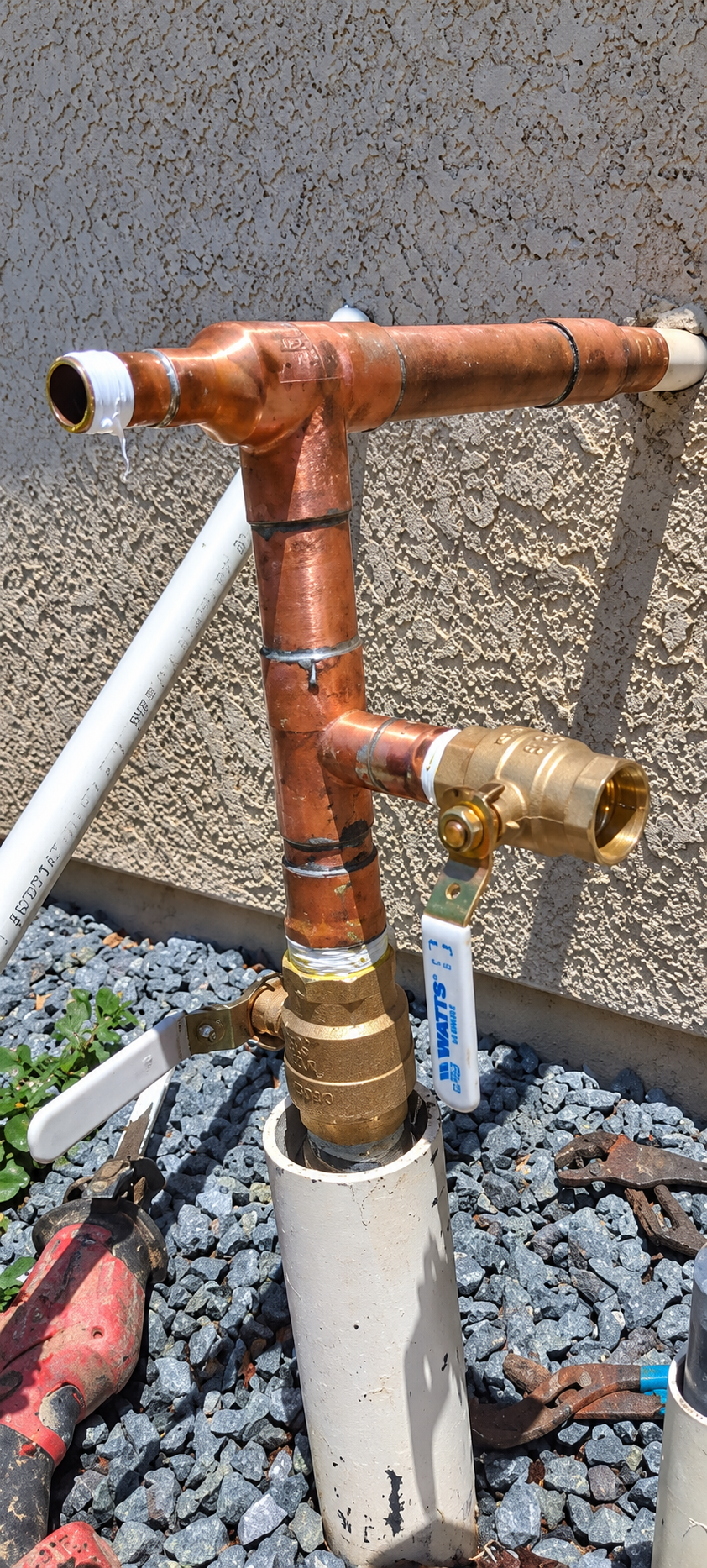 Rusty copper pipes and brass fittings on a white PVC base against gravel and stucco wall.