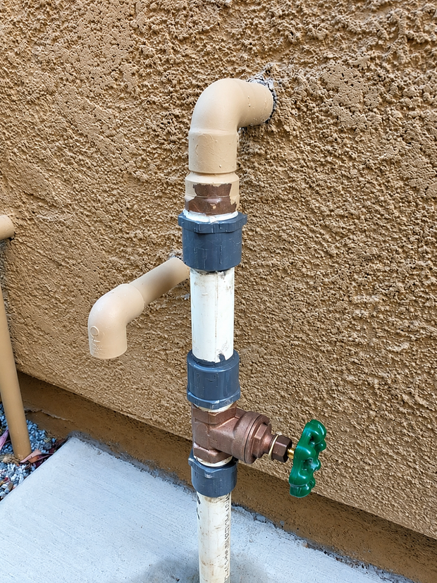 Outdoor white PVC pipes with blue couplings, an elbow joint, and a green valve against a stucco wall