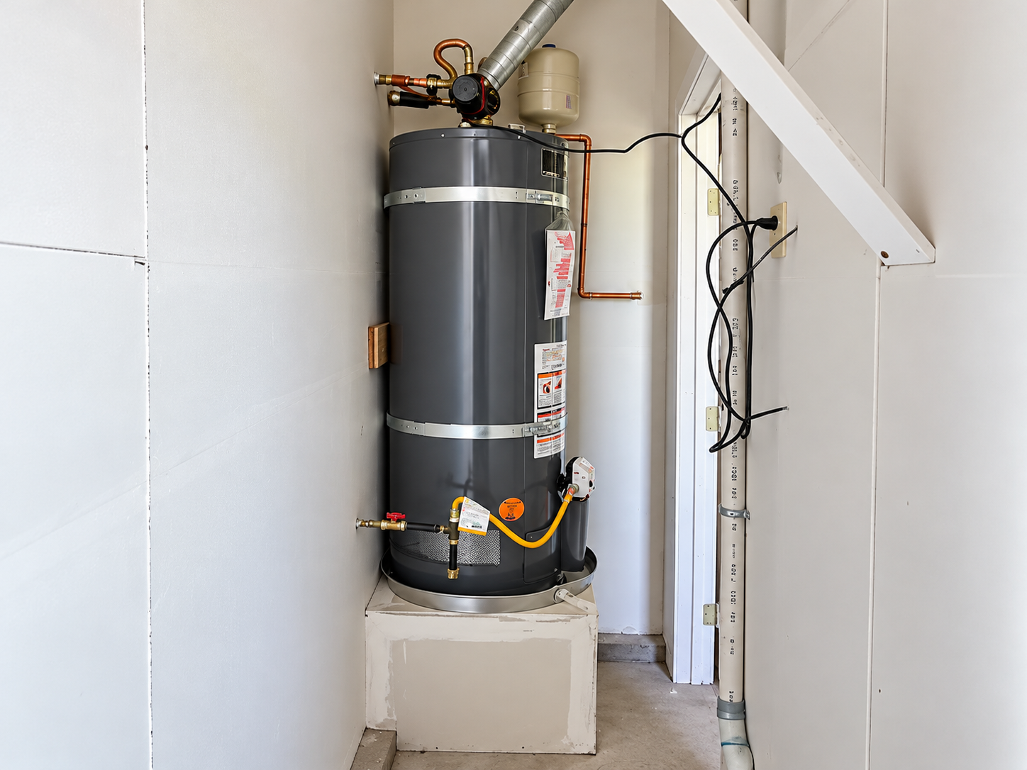 Water heater in a narrow utility closet with exposed pipes and wiring, atop a white pedestal
