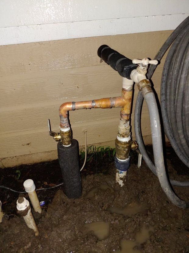 Copper pipes and outdoor plumbing valves against a wall, with hoses and muddy ground nearby