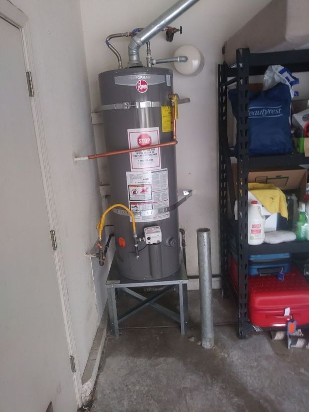 Water heater in a garage corner, strapped to a stand beside shelving and cleaning supplies.