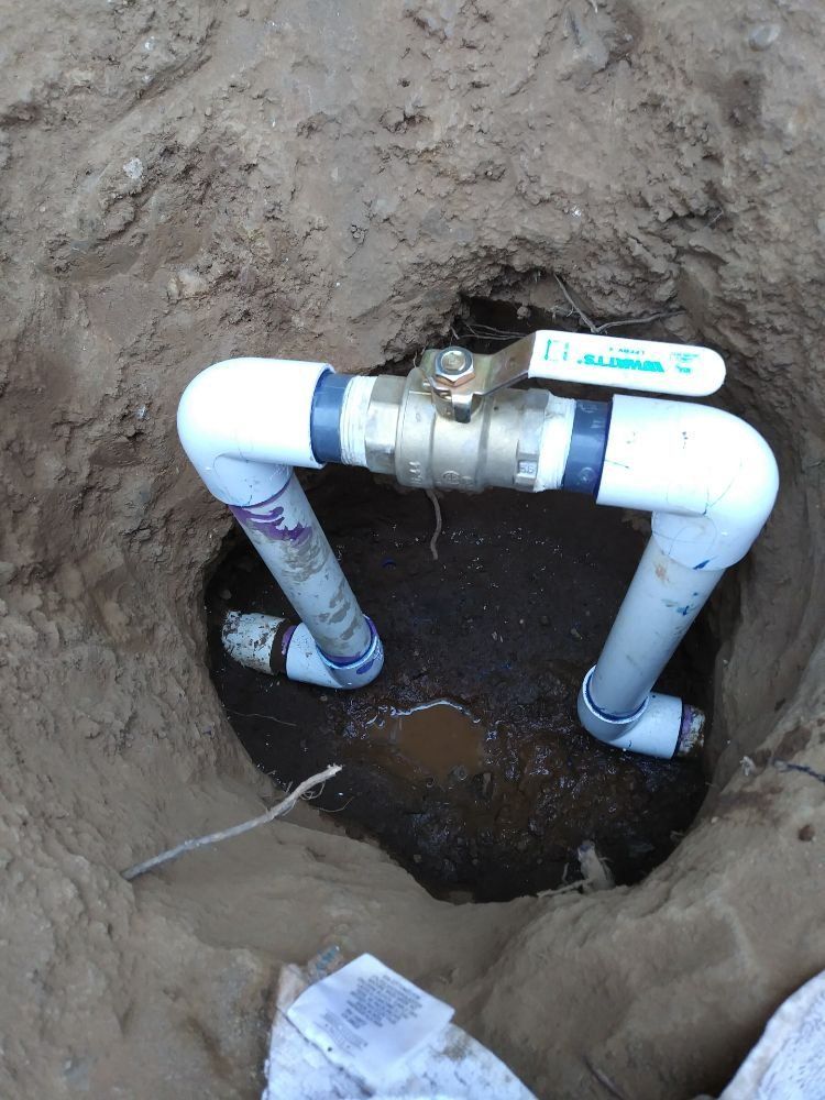 Exposed PVC plumbing pipes in a dirt trench with standing water at the bottom