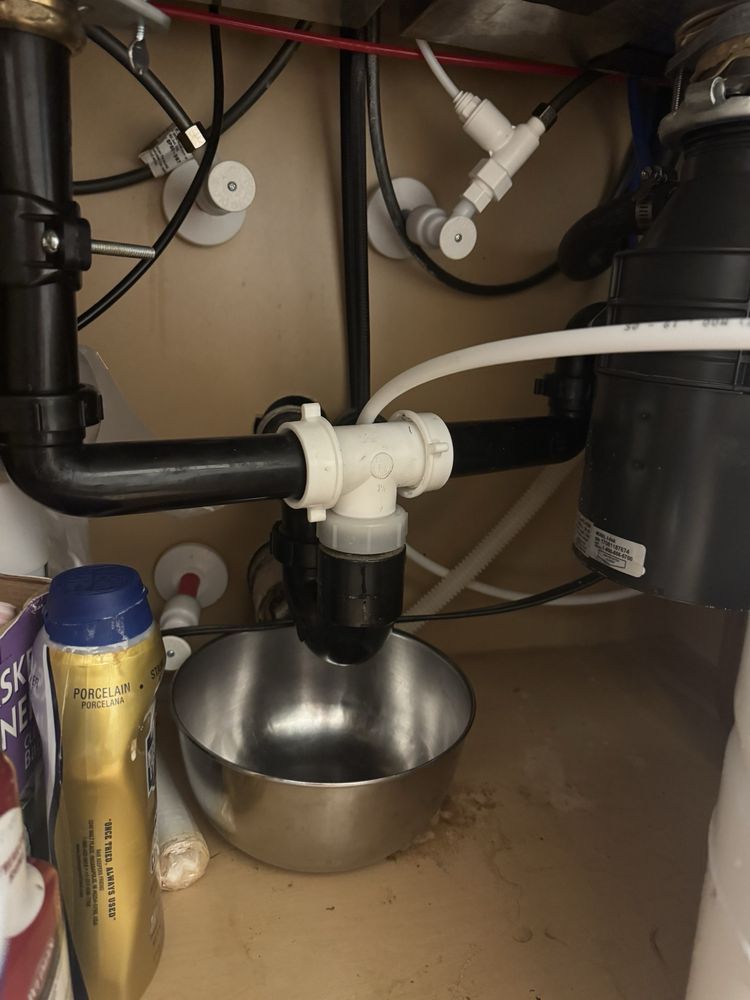 Under-sink plumbing with black and white drain pipes and a metal bowl on the cabinet floor