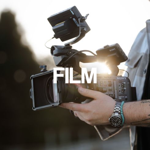 En person som holder et profesjonelt videokamera, med «FILM» over. Bakgrunnsbelyst sol.