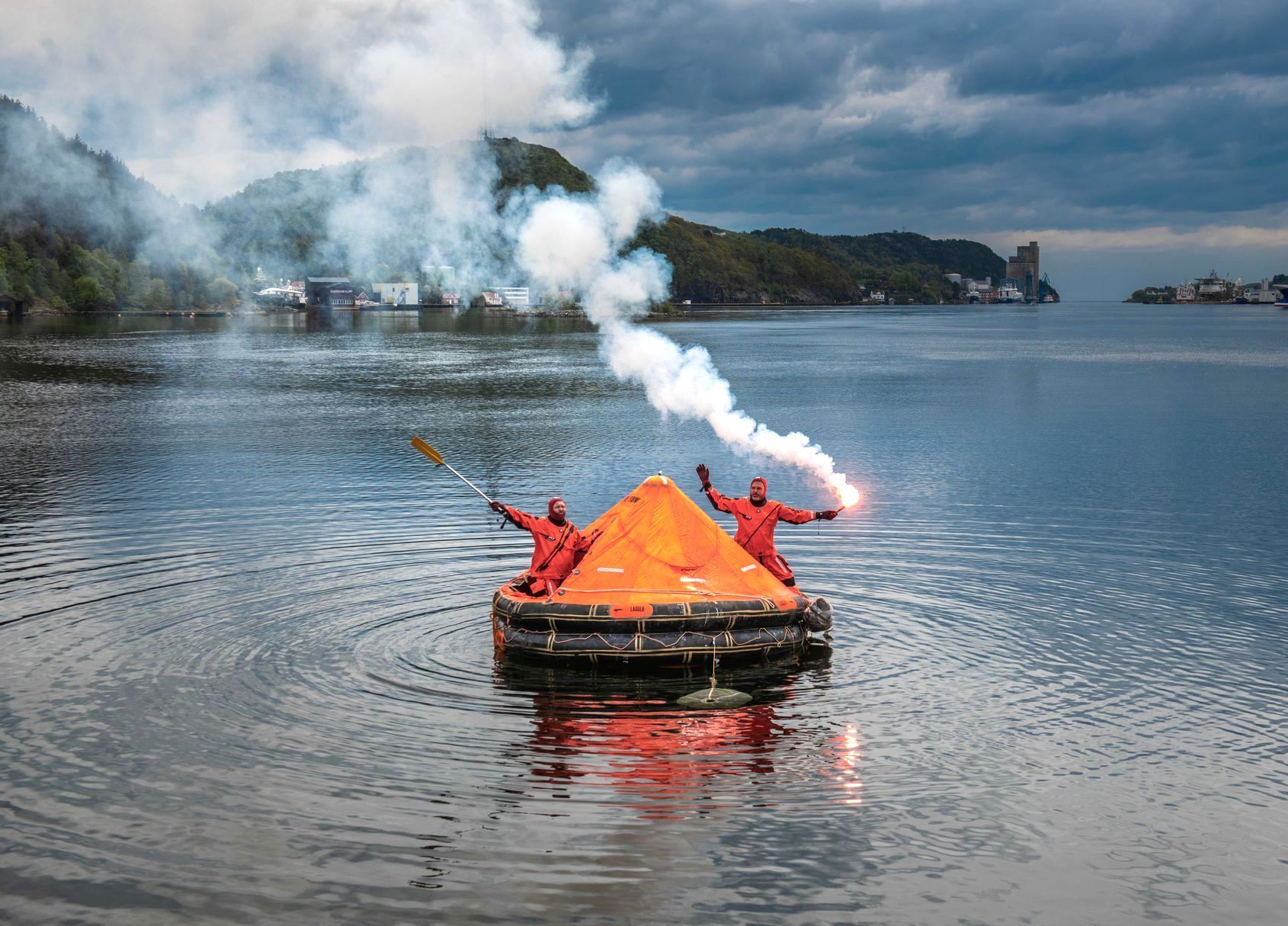 To personer i oransje overlevelsesdrakter på en oppblåsbar flåte på vannet, som aktiverer signalbluss som avgir hvit røyk mot en overskyet himmel.
