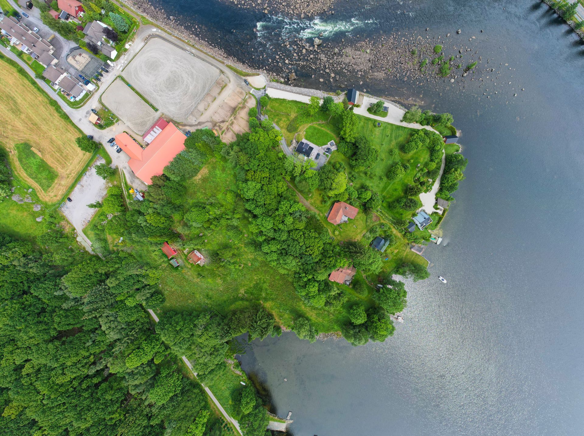 Luftfoto av en eiendom ved innsjøen med bygninger, grøntområder og et vann. En grusbane og hus er synlige.