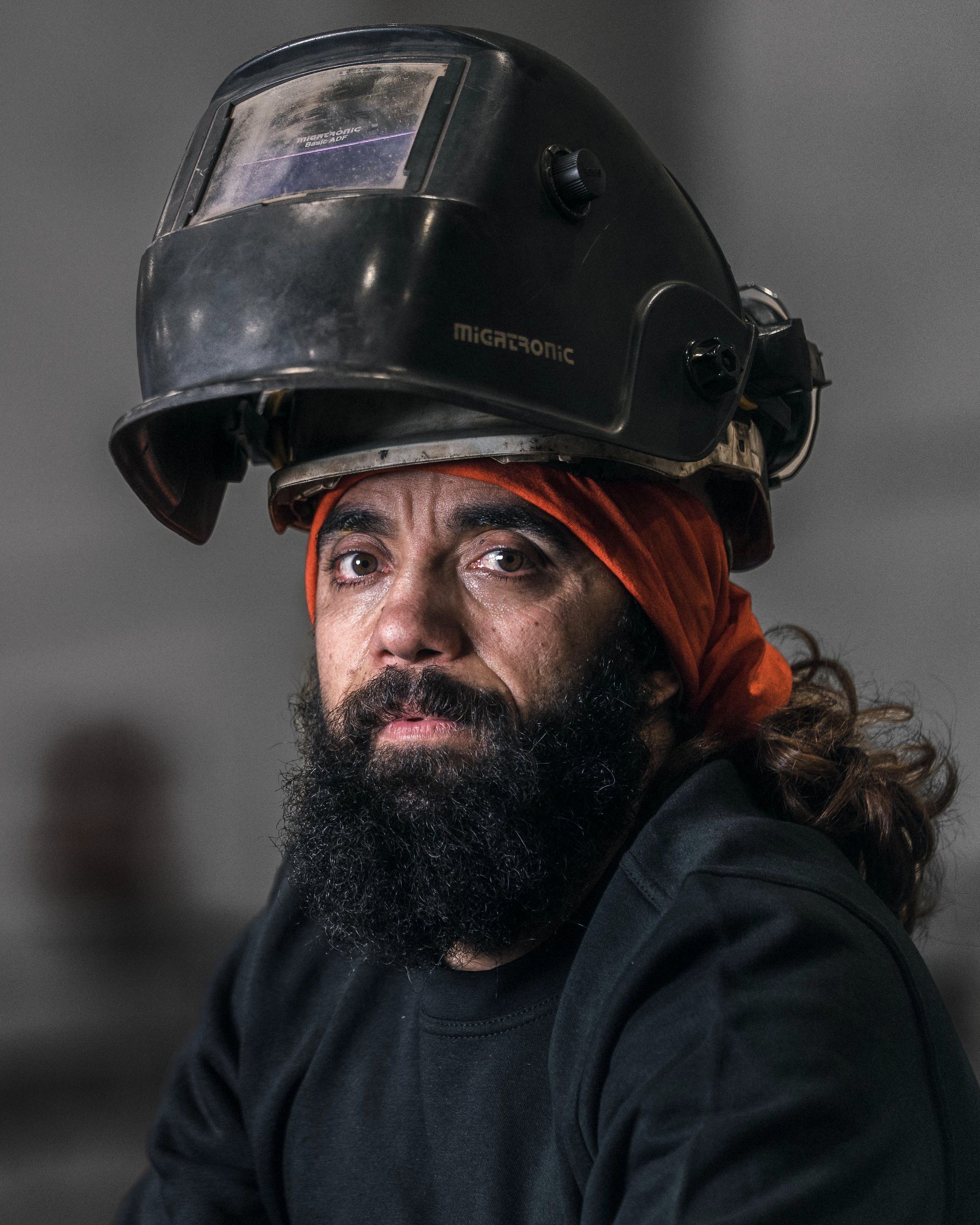 Sveiser med svart hjelm og oransje hodeskjerf, ser på betrakteren med et fokusert uttrykk.