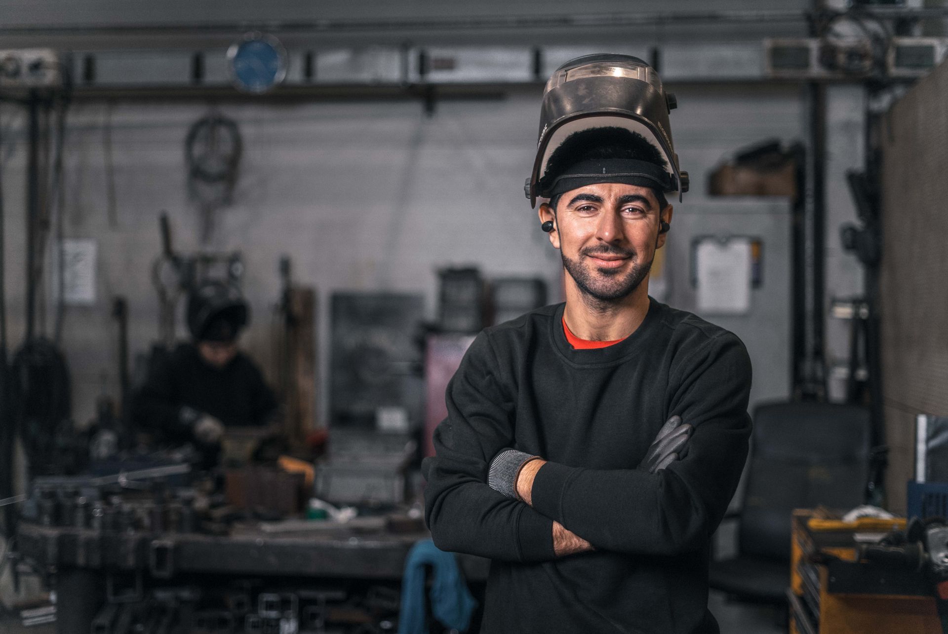Smilende sveiser i et verksted med armene i kors og sveisehjelm på hodet. En annen sveiser jobber i bakgrunnen.