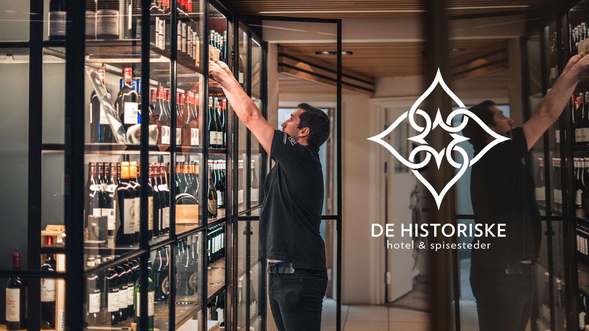 Mann rekker etter en flaske i en vinkjeller på De Historiske. Mørke tre- og metallhyller viser frem mange vinflasker.