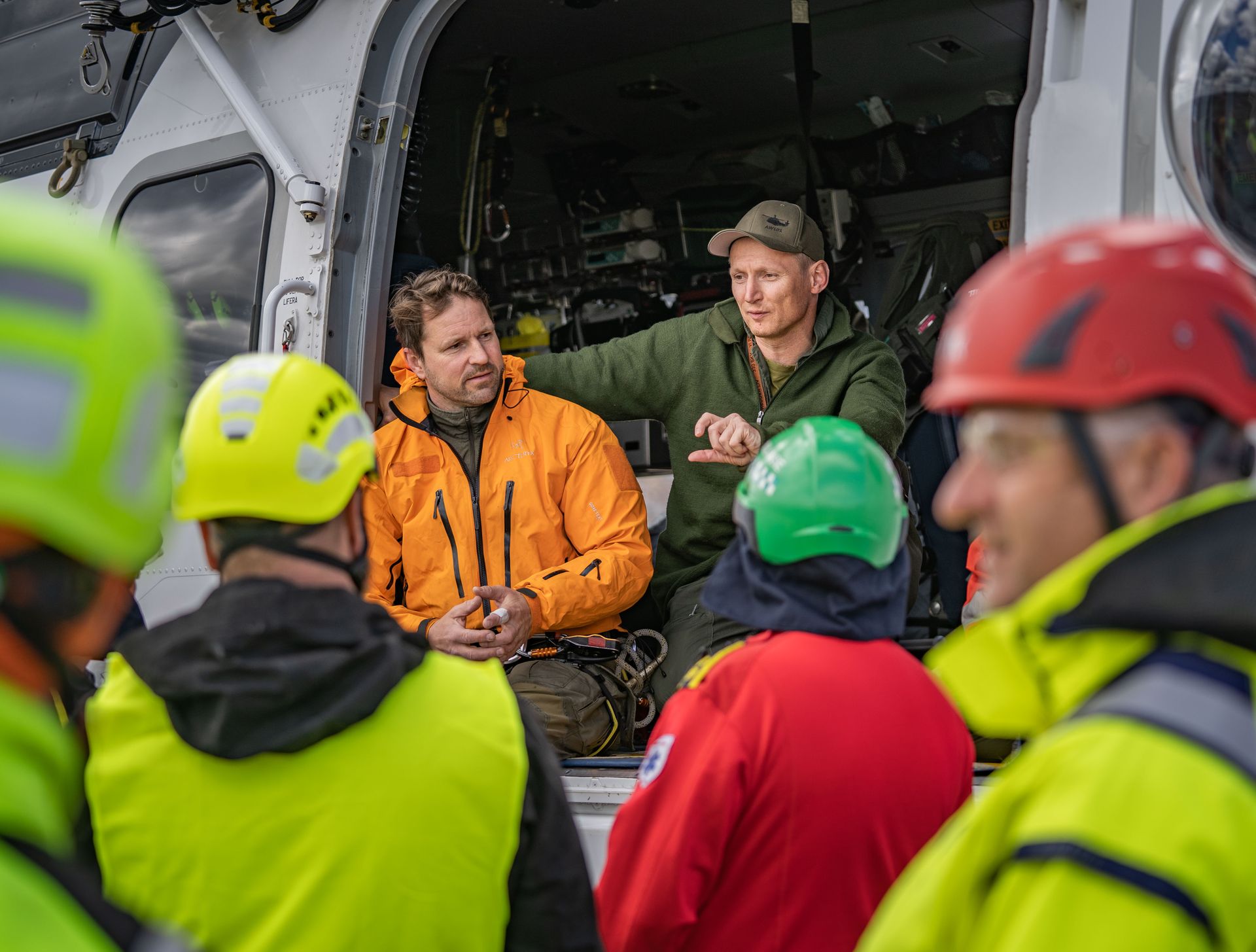 En redningsmannskapsbriefing nær et helikopter. To menn inne i helikopteret, én peker. Andre teammedlemmer i lyst sikkerhetsutstyr.