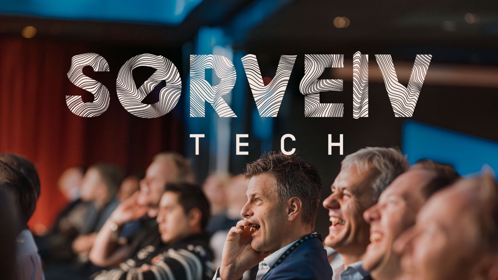Publikum på Søveiv Tech-arrangementet, for det meste hvite menn, lo. Arrangementsteksten «SØRVEIV TECH» i hvitt overlegg.