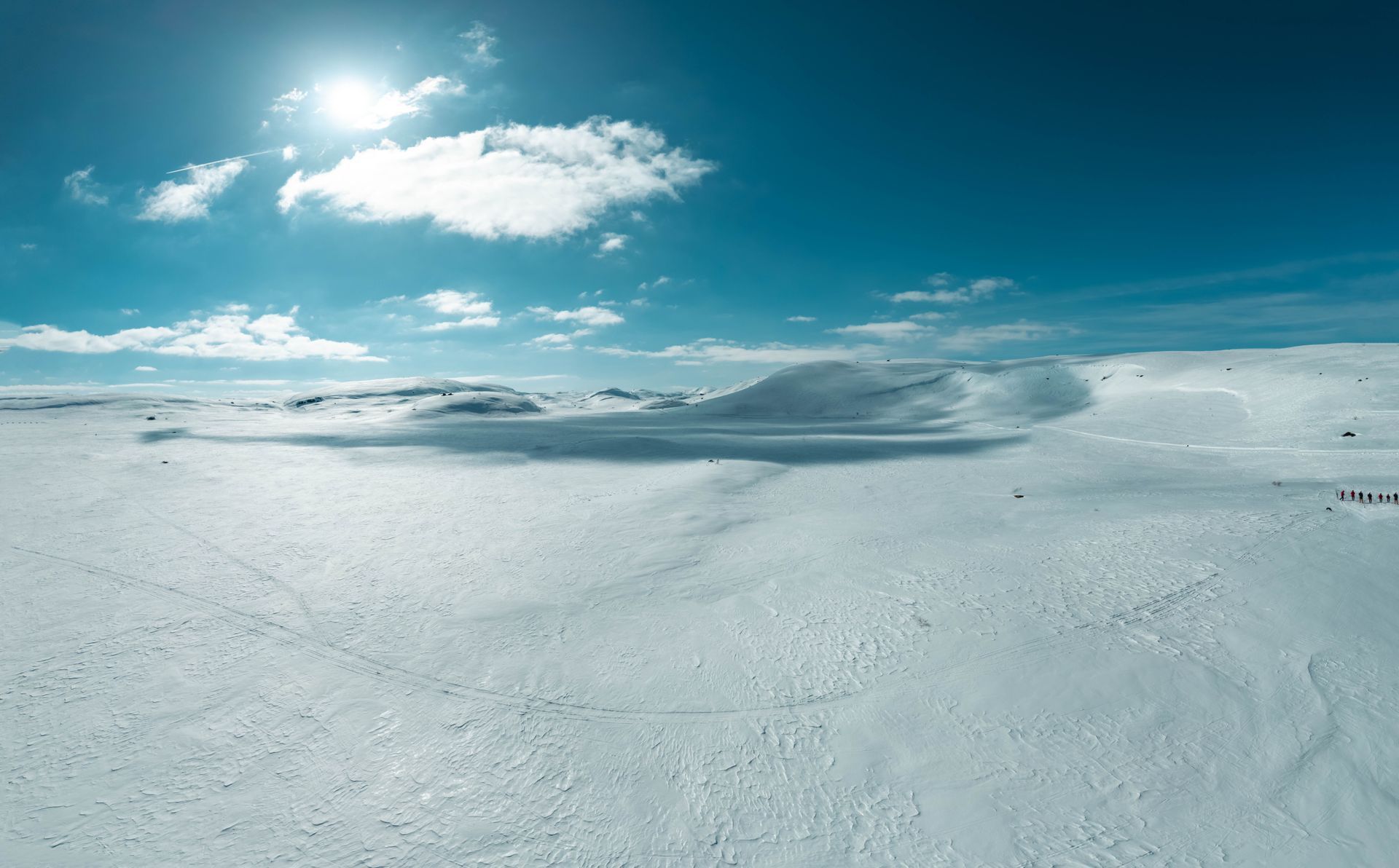 Et vidstrakt, snødekt landskap under en strålende sol og blå himmel, med noen få mørke skikkelser som ser små ut i det fjerne.