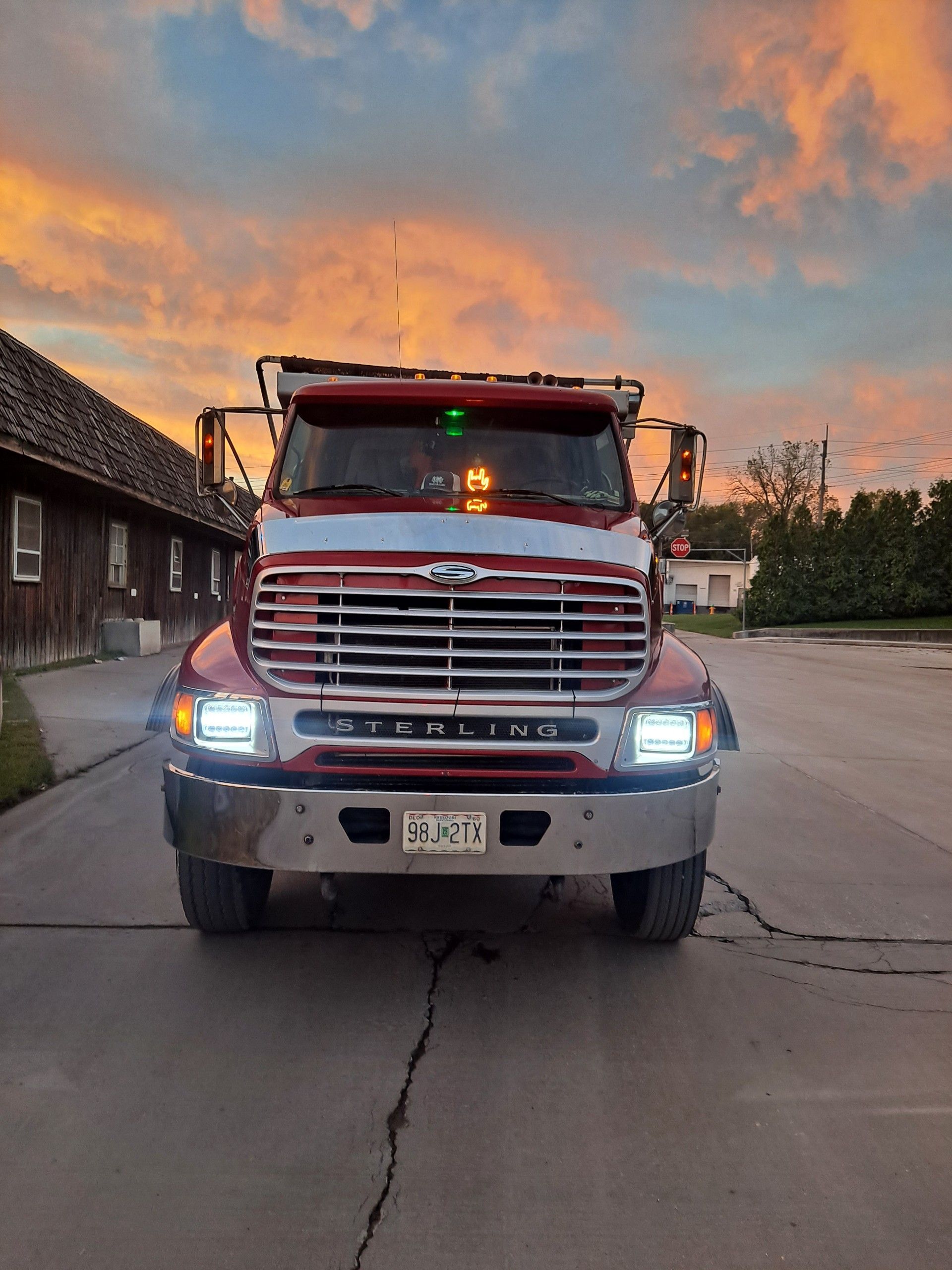 Red Sterling dump truck on cracked asphalt road, sunset sky in background.