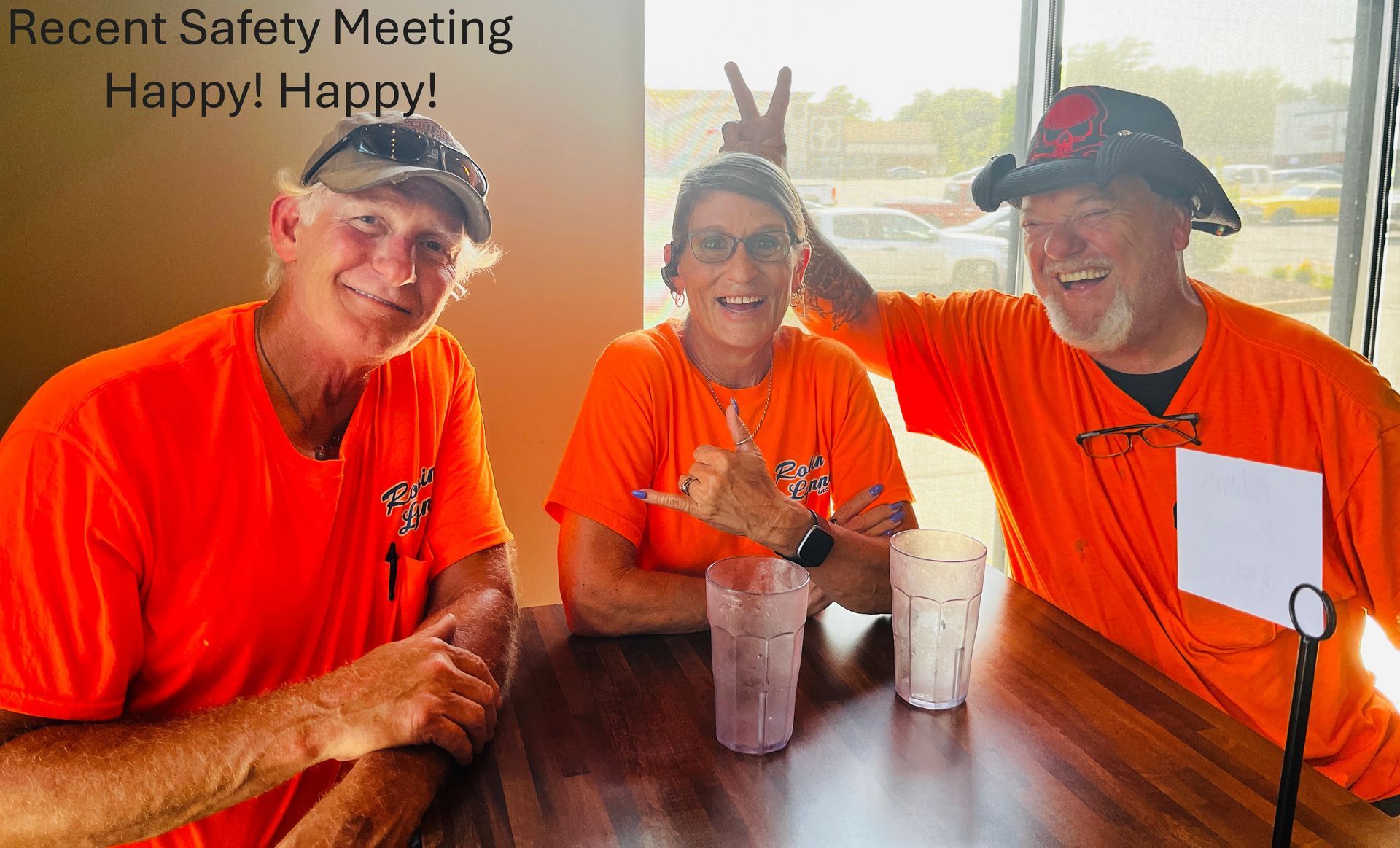 Three people in bright orange shirts smiling at a table, holding drinks at a Happy Halloween safety meeting.