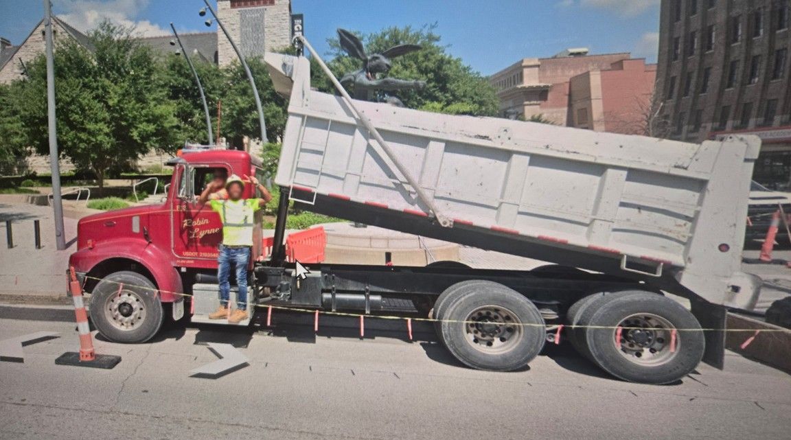 Red dump truck with raised bed; worker in vest stands near it, arms raised. Street setting.