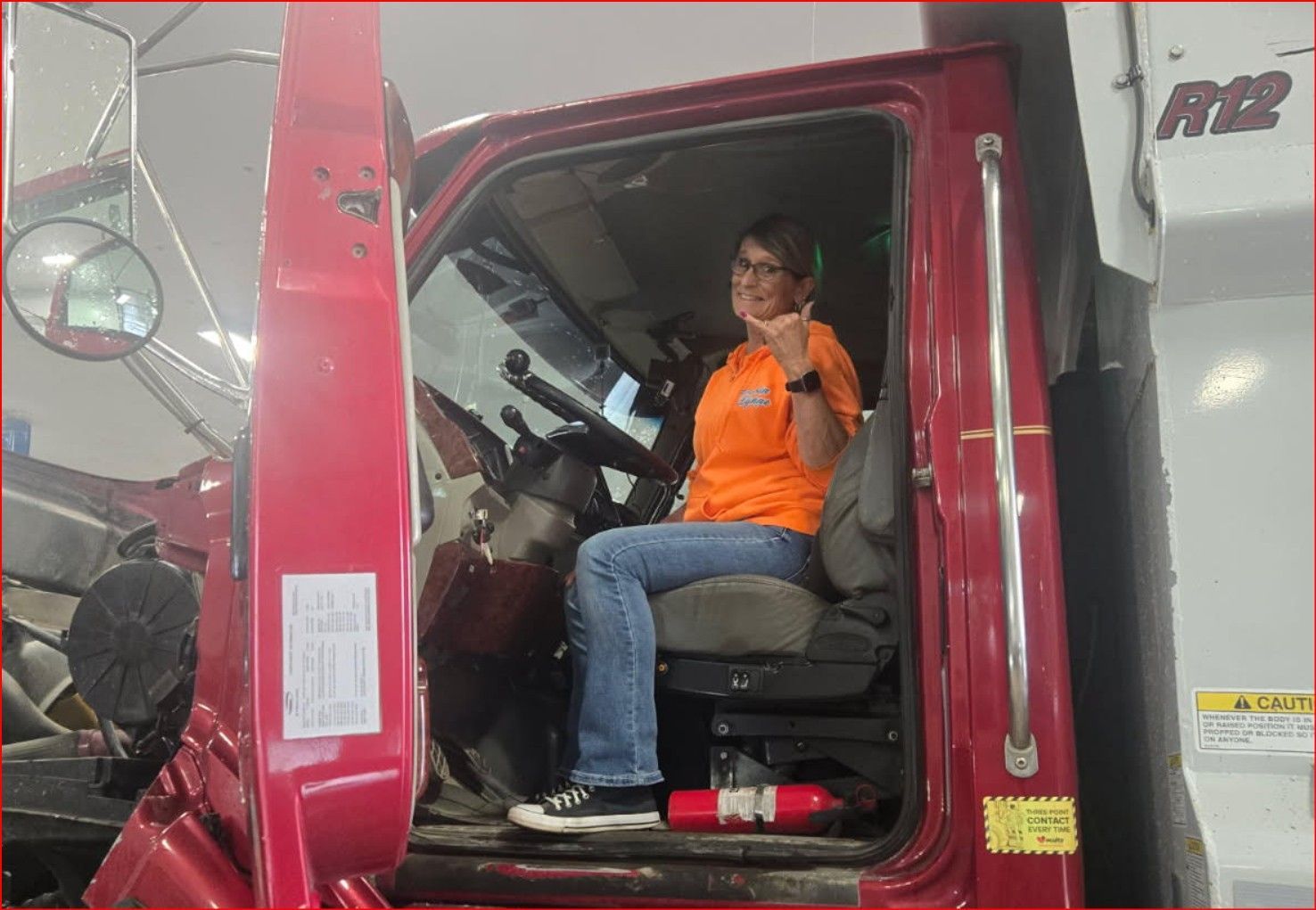 Person in orange shirt sitting in a red truck cab, smiling and waving.