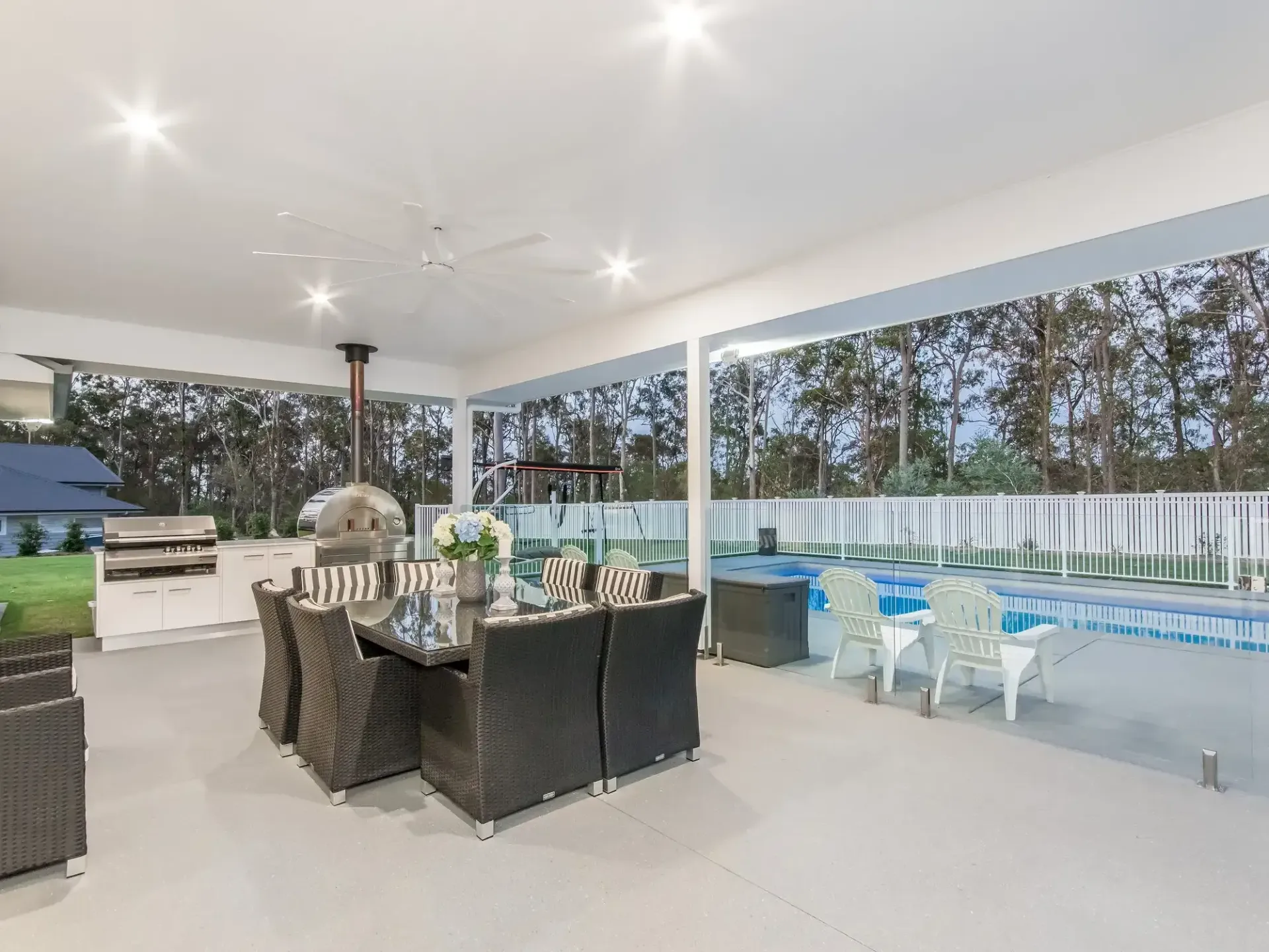 A modern outdoor covered patio with a dark wicker dining set, a pizza oven, and a glass-fenced pool in the background. — Michael Duncan Constructions Pty Ltd in Robina, QLD