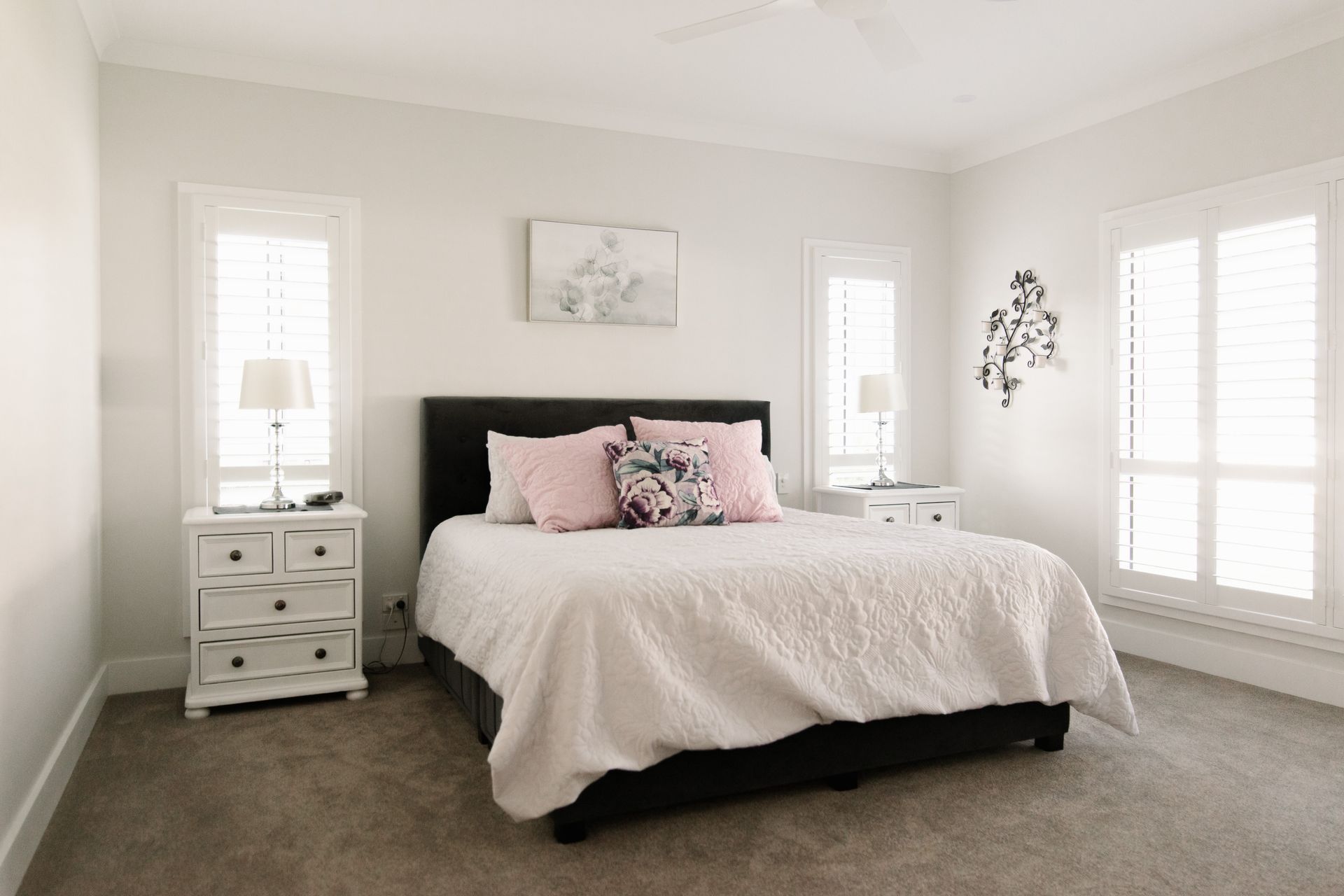 A bedroom with a dark bed frame, white textured quilt, pink pillows, two white nightstands with lamps, and plantation blinds. — Michael Duncan Constructions Pty Ltd in Burleigh Heads, QLD