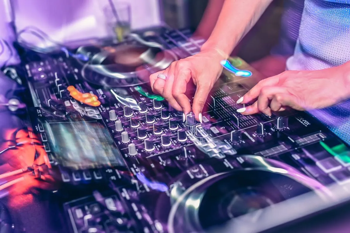 Hands on DJ mixer, adjusting controls with focus on lights and equipment in a dimly lit setting.