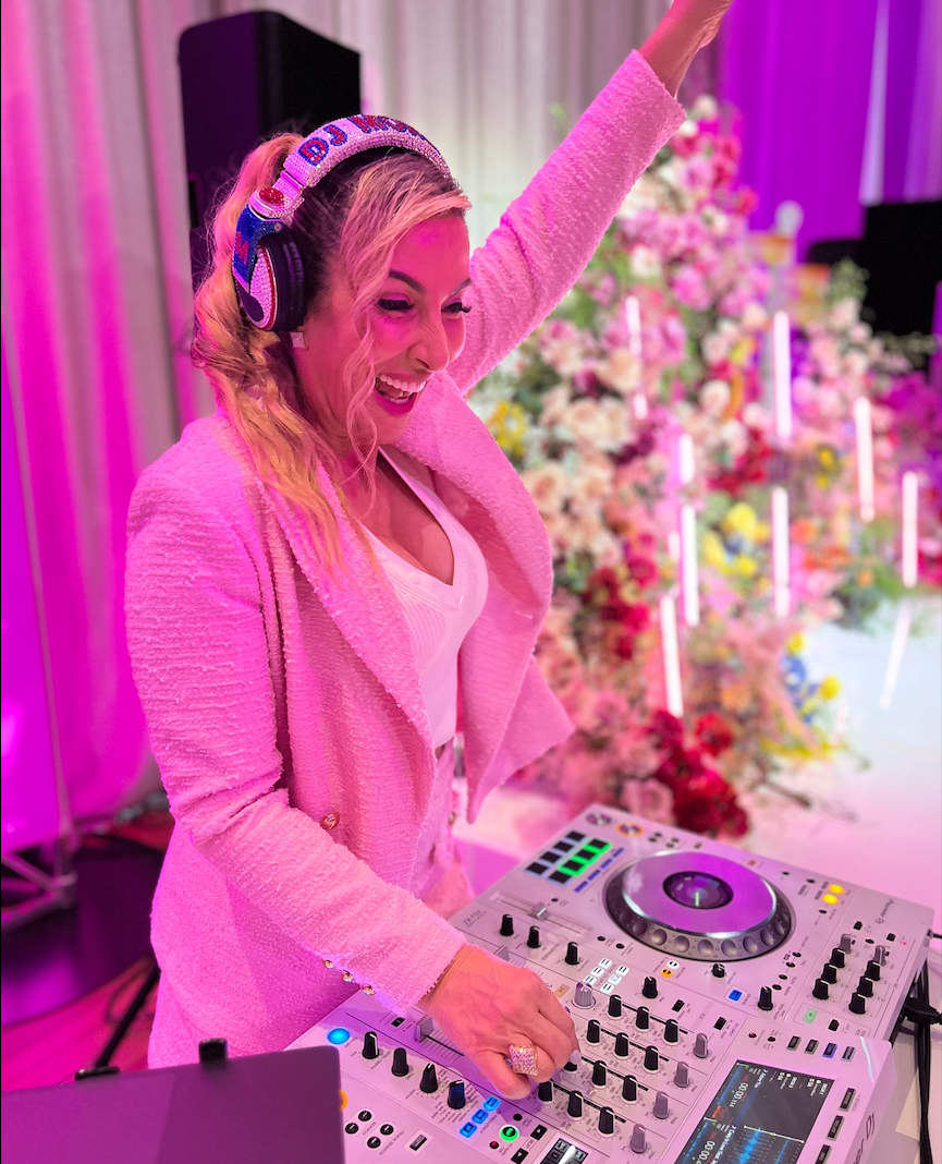 Blonde woman DJ in pink suit with raised arm, smiling, at a lit event with floral backdrop.