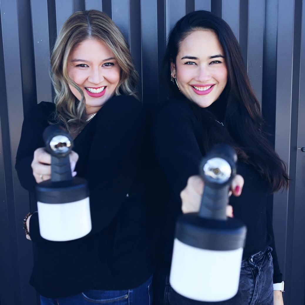 Two smiling women posing with spray bottles in front of a gray wall
