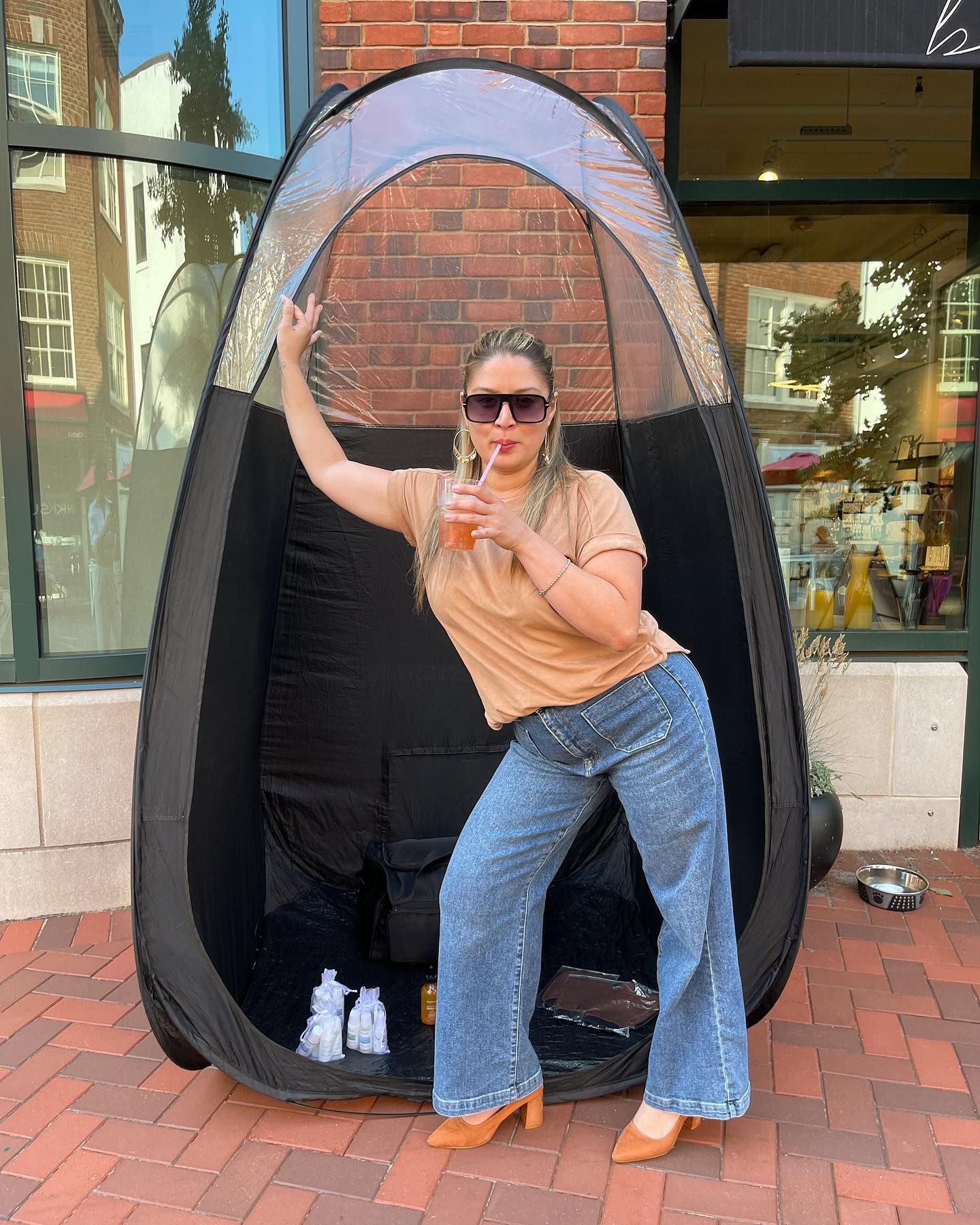 Woman posing beside a black outdoor pod, wearing sunglasses, peach top, and jeans on a brick sidewalk.