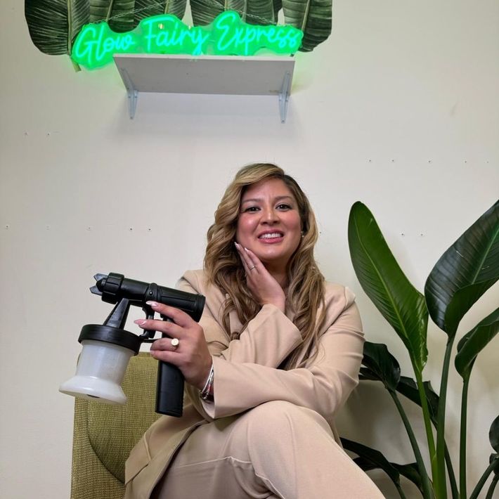 Woman sitting indoors holding a spray gun, smiling beside a green neon sign and potted plant.