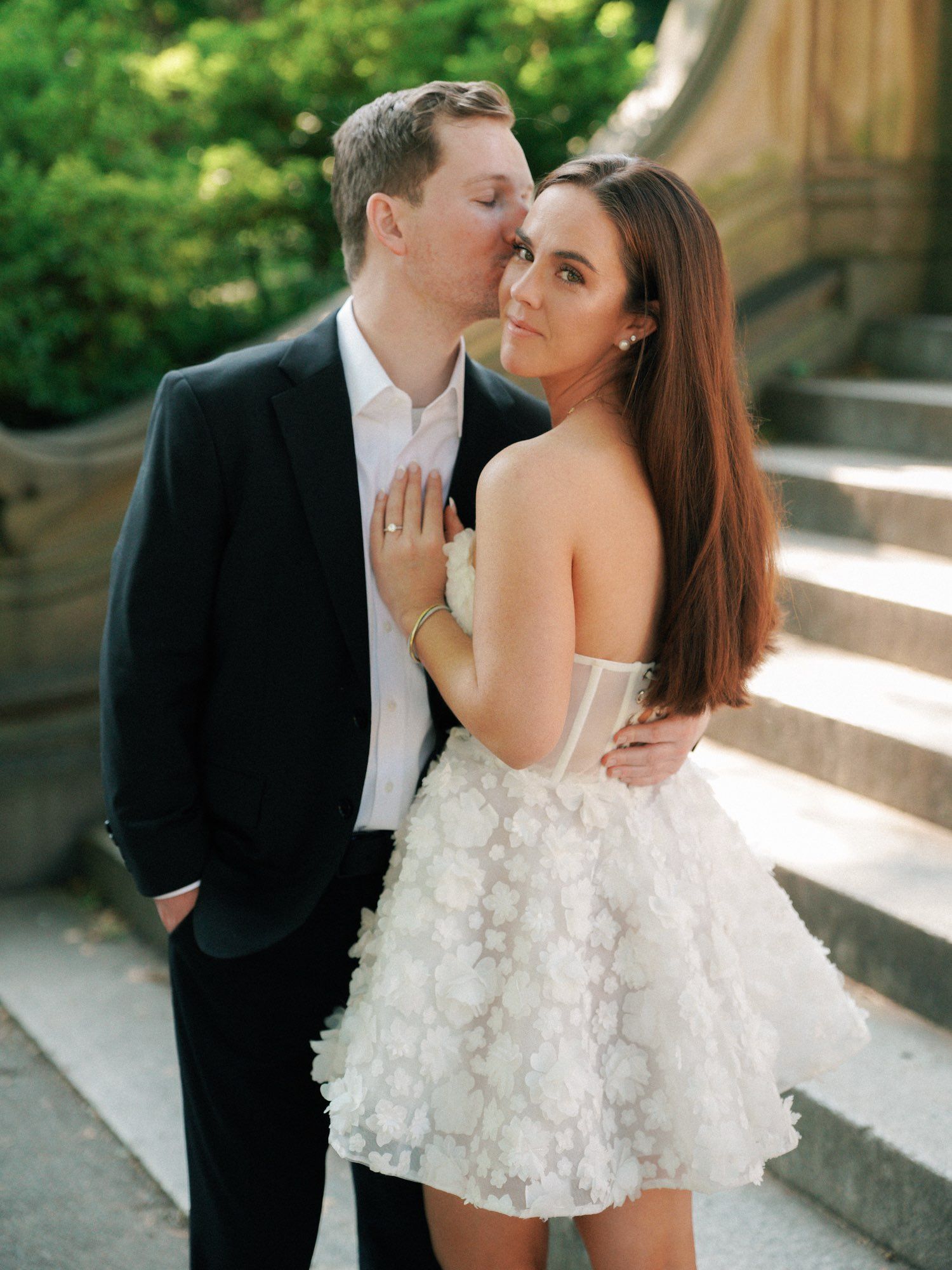 Couple in formal attire sharing a kiss outdoors, with the woman in a white dress and the man in a black suit.
