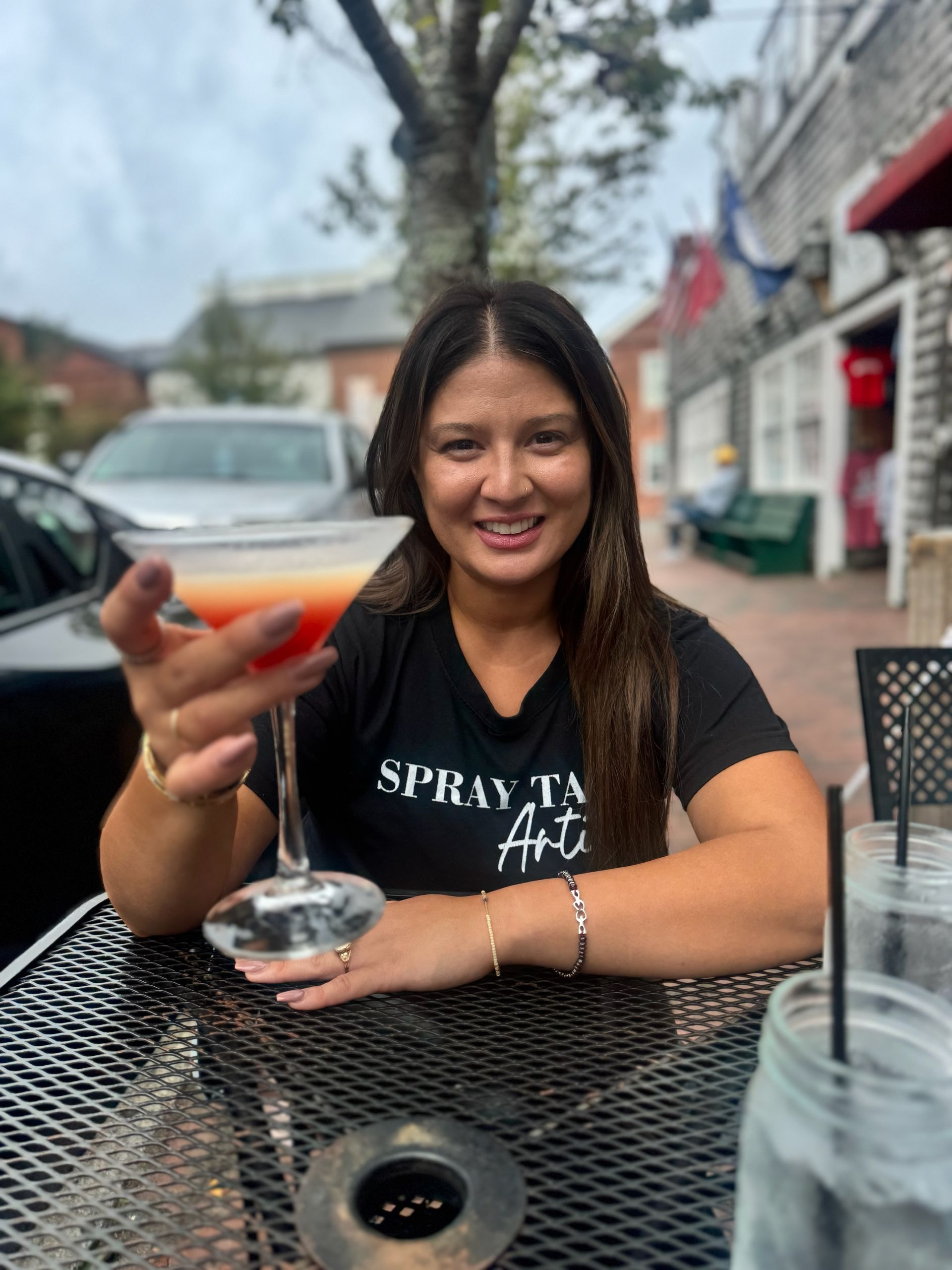 Woman smiling at an outdoor café, holding a drink and sitting at a black metal table.