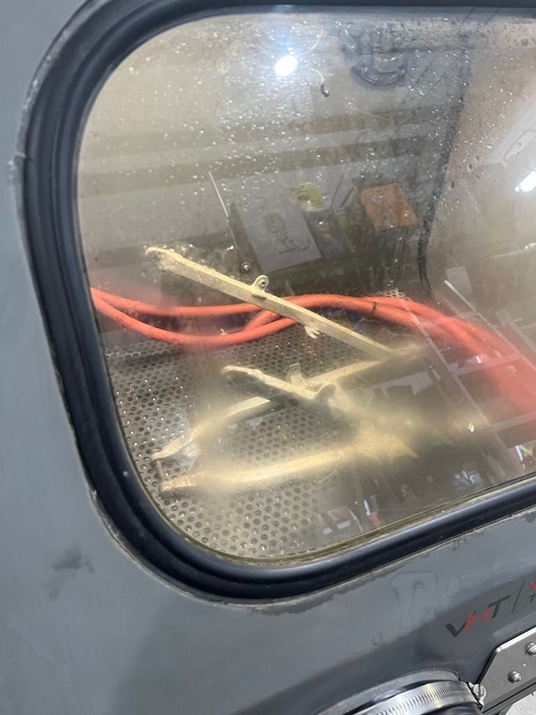 A metal frame and red cables inside a sandblasting cabinet with a glass view window. A metal frame and red cables inside a sandblasting cabinet with a glass view window.