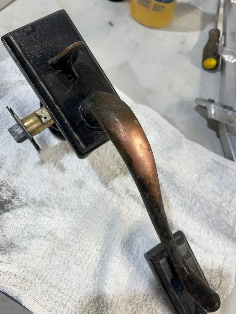 A rustic black door handle with a curved, tarnished copper-toned grip lying on a white cloth surface. A rustic black door handle with a curved, tarnished copper-toned grip lying on a white cloth surface.
