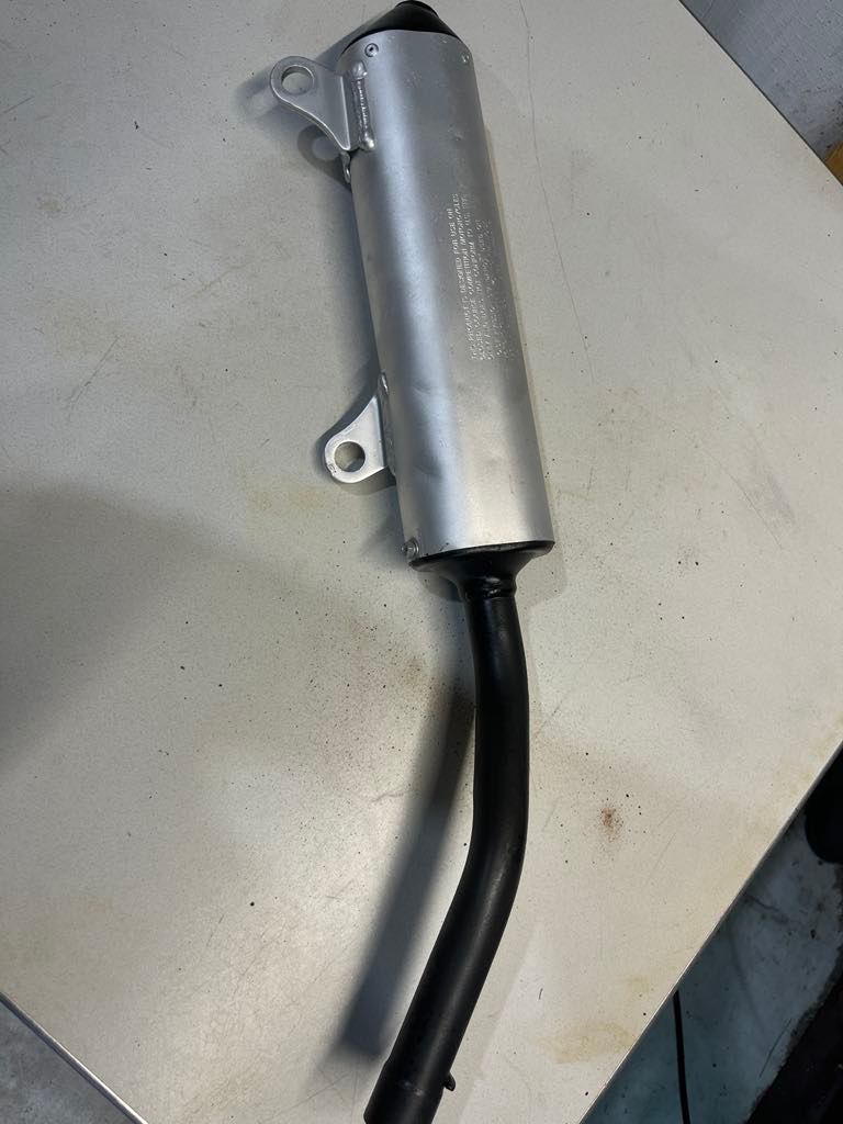 A motorcycle exhaust muffler with a silver cylindrical body and a curved black pipe, viewed from above on a flat surface. A motorcycle exhaust muffler with a silver cylindrical body and a curved black pipe, viewed from above on a flat surface.