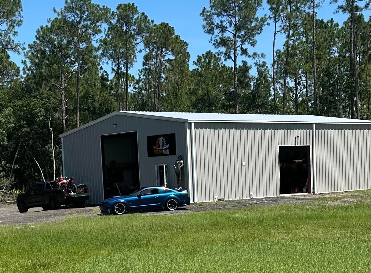 A blue sports car parked in front of a large metal warehouse building surrounded by a dense forest on a sunny day. A blue sports car parked in front of a large metal warehouse building surrounded by a dense forest on a sunny day.