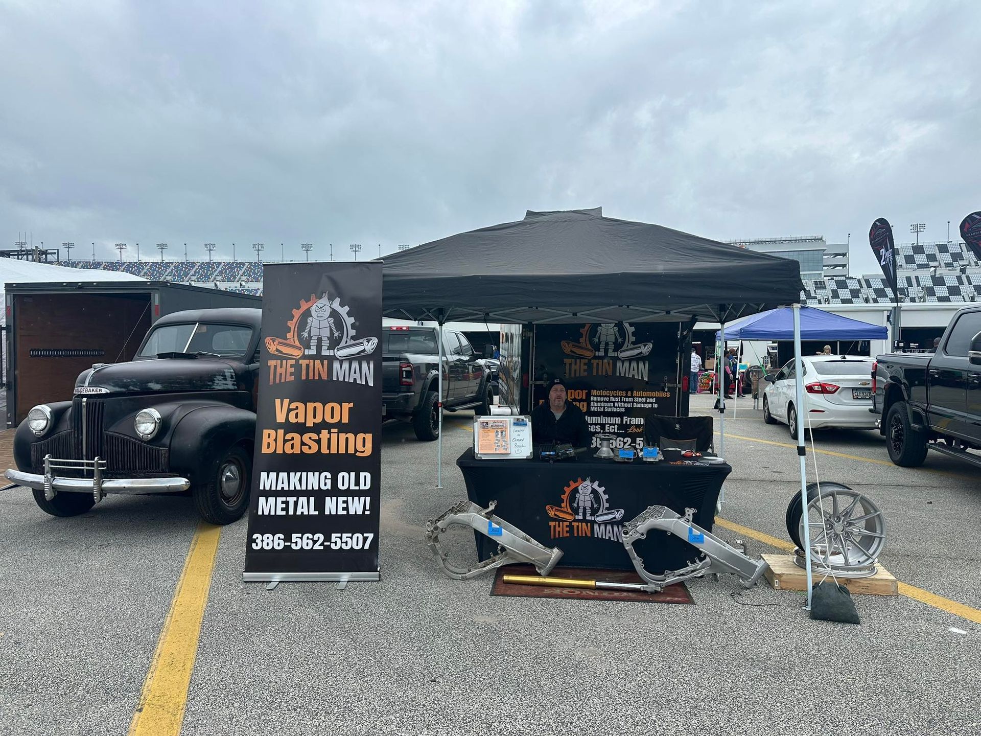 A The Tin Man Vapor Blasting booth with a vintage black car and metal parts on display at an outdoor event. A The Tin Man Vapor Blasting booth with a vintage black car and metal parts on display at an outdoor event.
