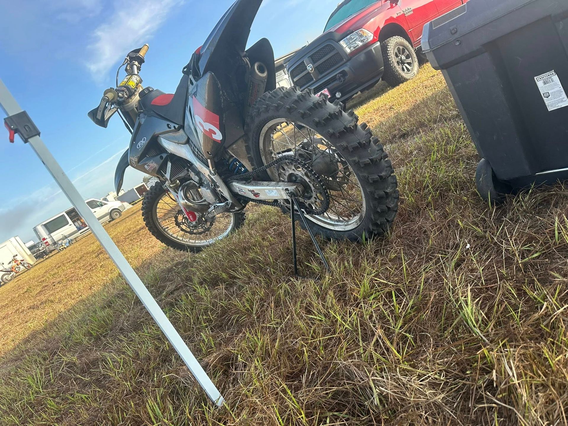 A black and red dirt bike parked on grass near a red truck and a grey storage bin under a clear sky. A black and red dirt bike parked on grass near a red truck and a grey storage bin under a clear sky.