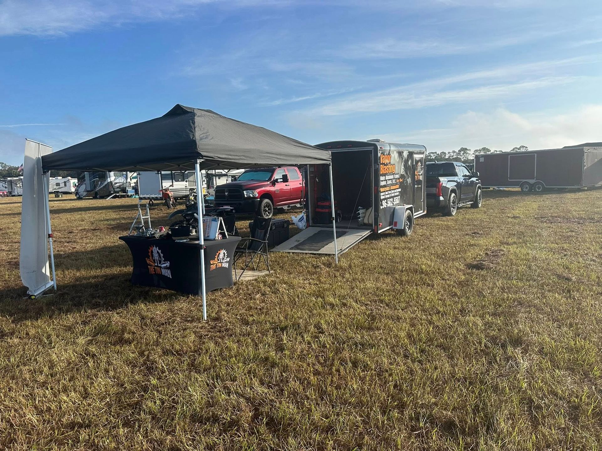 A black canopy tent with a branded table sits in a field next to an open equipment trailer and a red pickup truck. A black canopy tent with a branded table sits in a field next to an open equipment trailer and a red pickup truck.