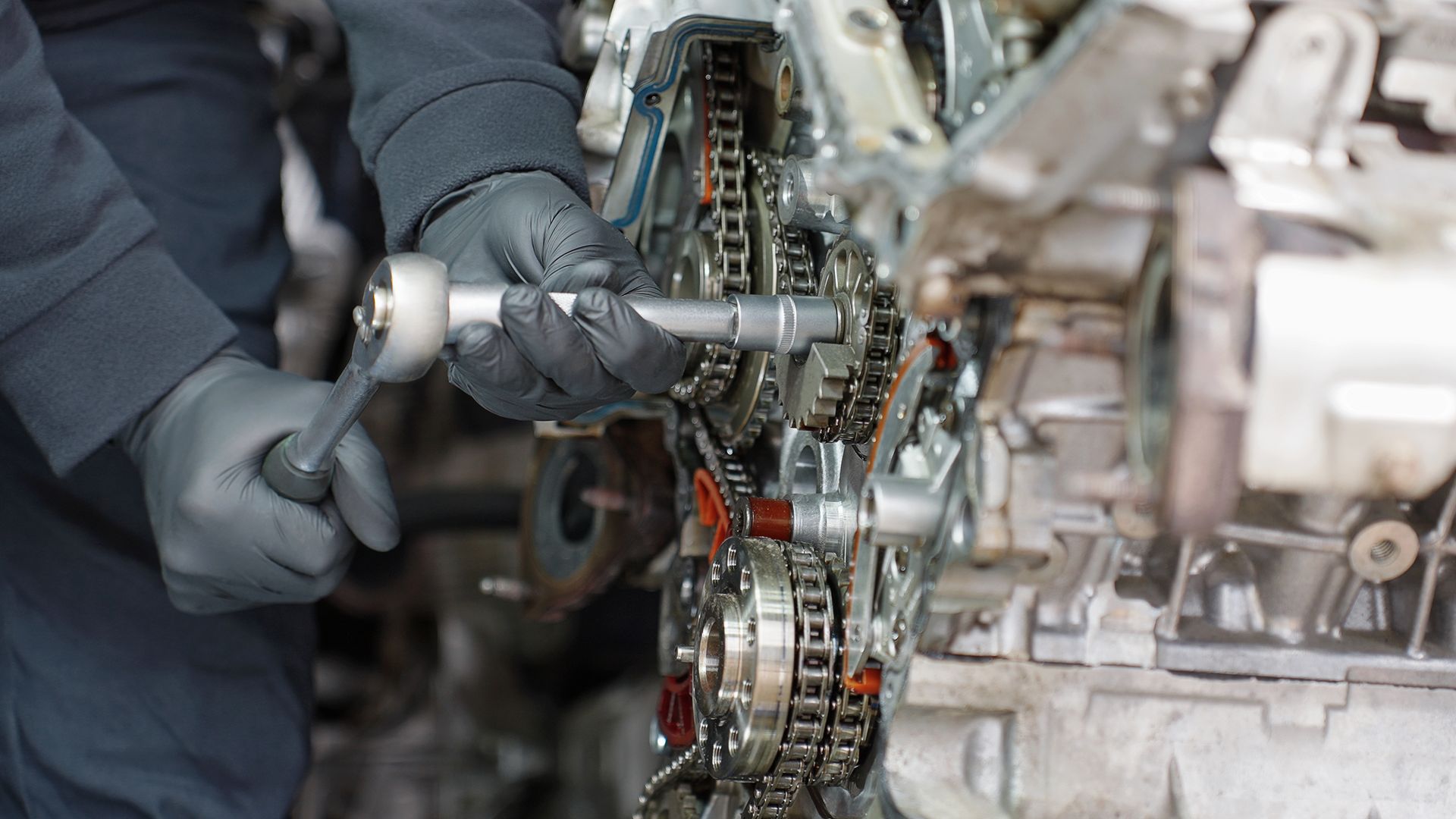 A mechanic wearing black gloves uses a ratchet tool to work on the internal timing chain mechanism of an engine. A mechanic wearing black gloves uses a ratchet tool to work on the internal timing chain mechanism of an engine.