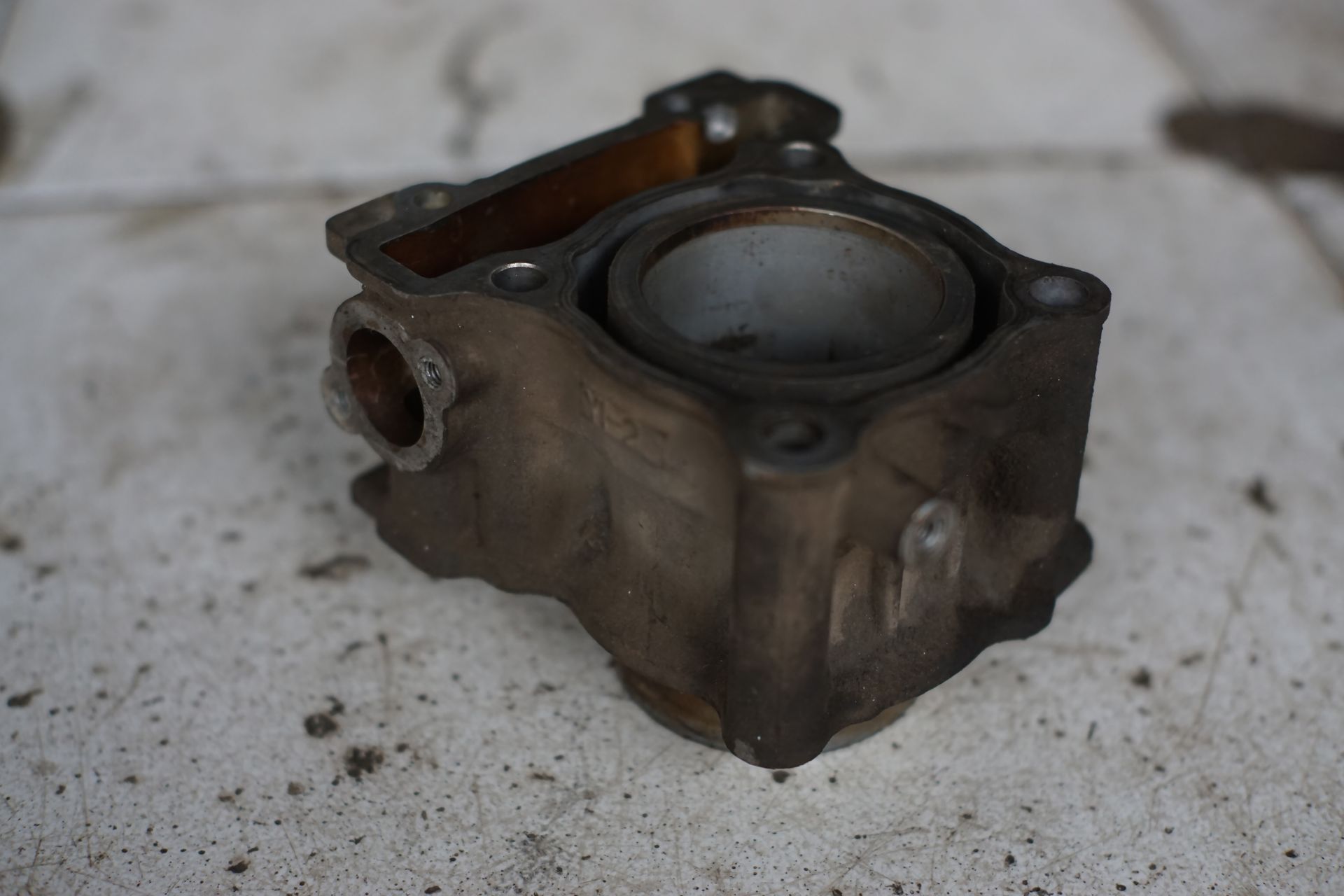 A used, dirty motorcycle engine cylinder block sitting on a stained concrete surface. A used, dirty motorcycle engine cylinder block sitting on a stained concrete surface.