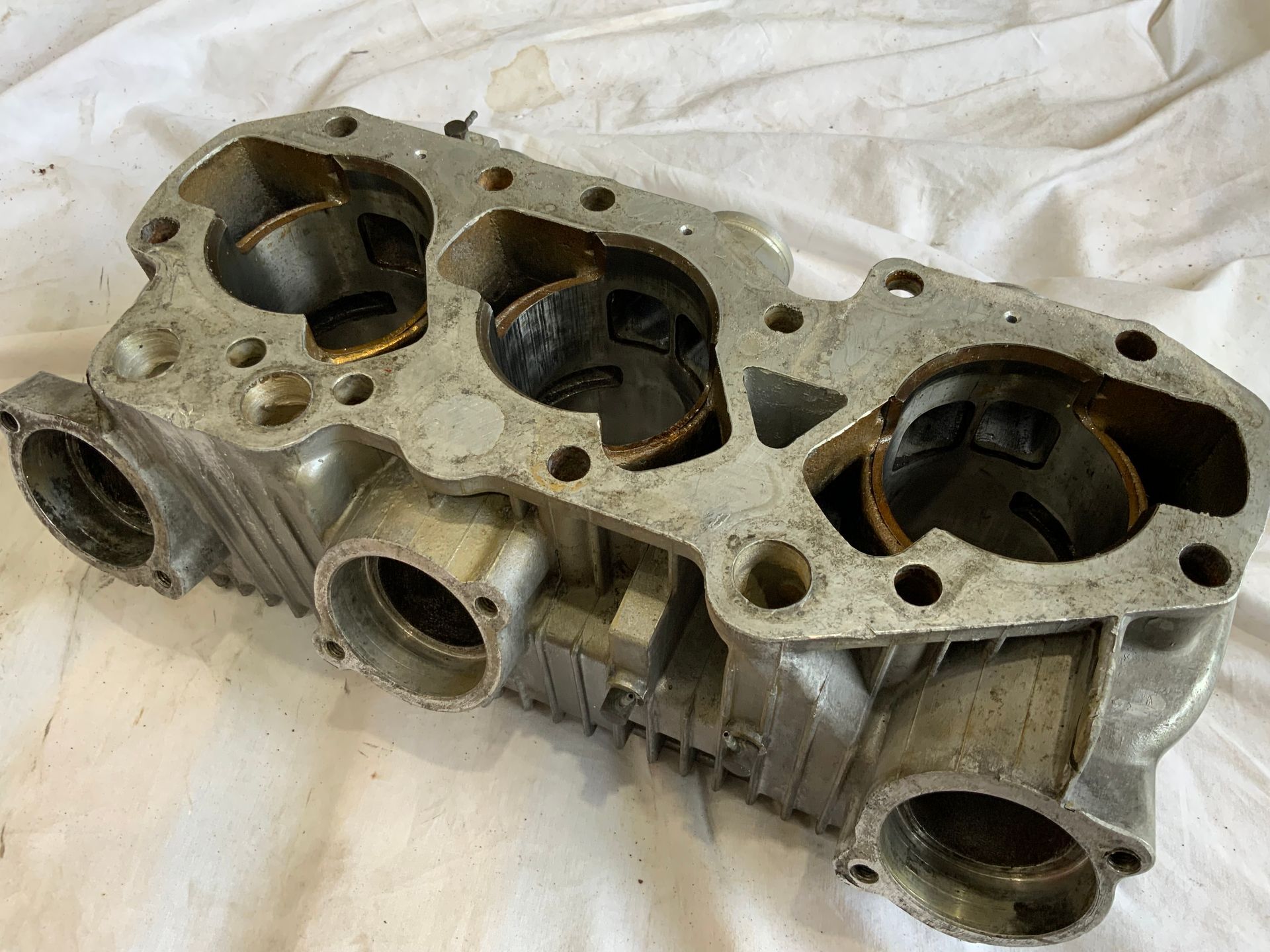 A weathered, three-cylinder engine block component made of cast metal, shown from an elevated, angled perspective. A weathered, three-cylinder engine block component made of cast metal, shown from an elevated, angled perspective.