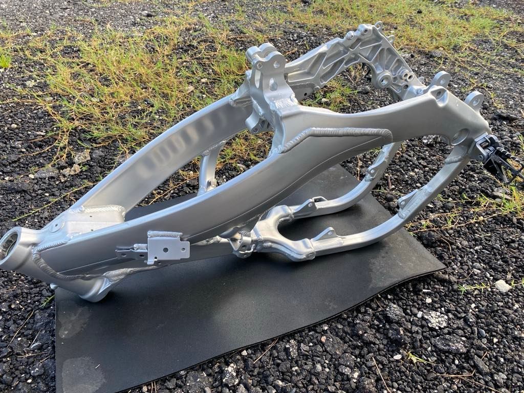 A silver aluminum motorcycle frame sitting on a black mat outdoors on a gravel surface. A silver aluminum motorcycle frame sitting on a black mat outdoors on a gravel surface.