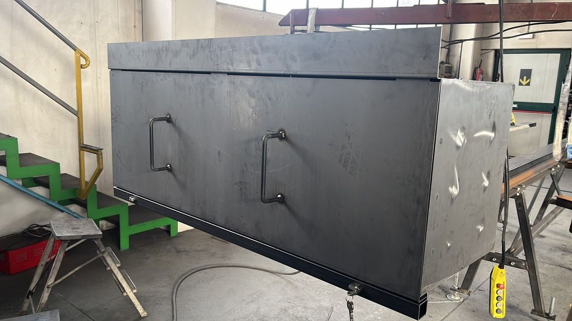 Grande scatola o serbatoio industriale in metallo nero in un'officina, con maniglie e scale verdi accanto.