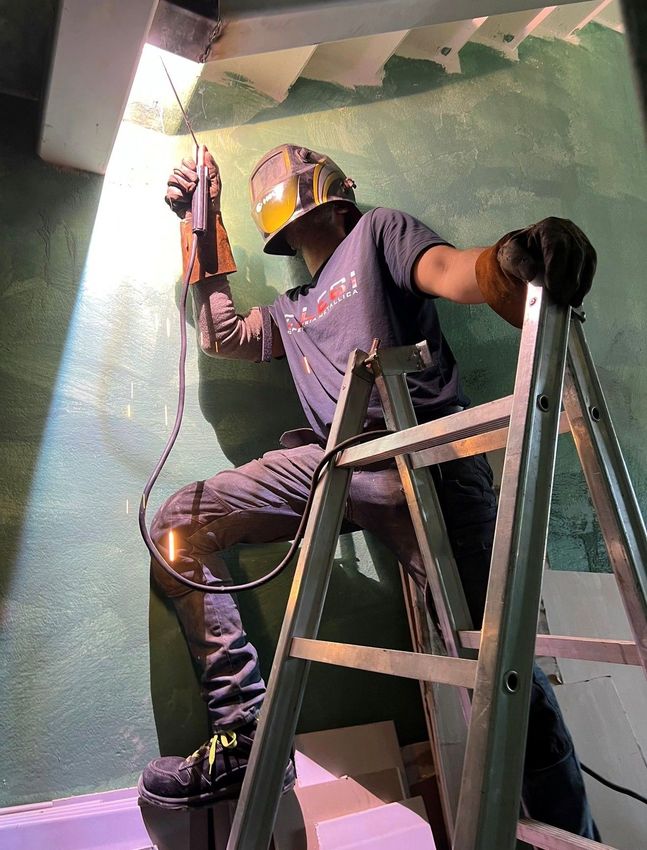 Una persona su una scala sta dipingendo una parete verde all'interno di un edificio, indossando un casco e tenendo in mano un rullo per dipingere.