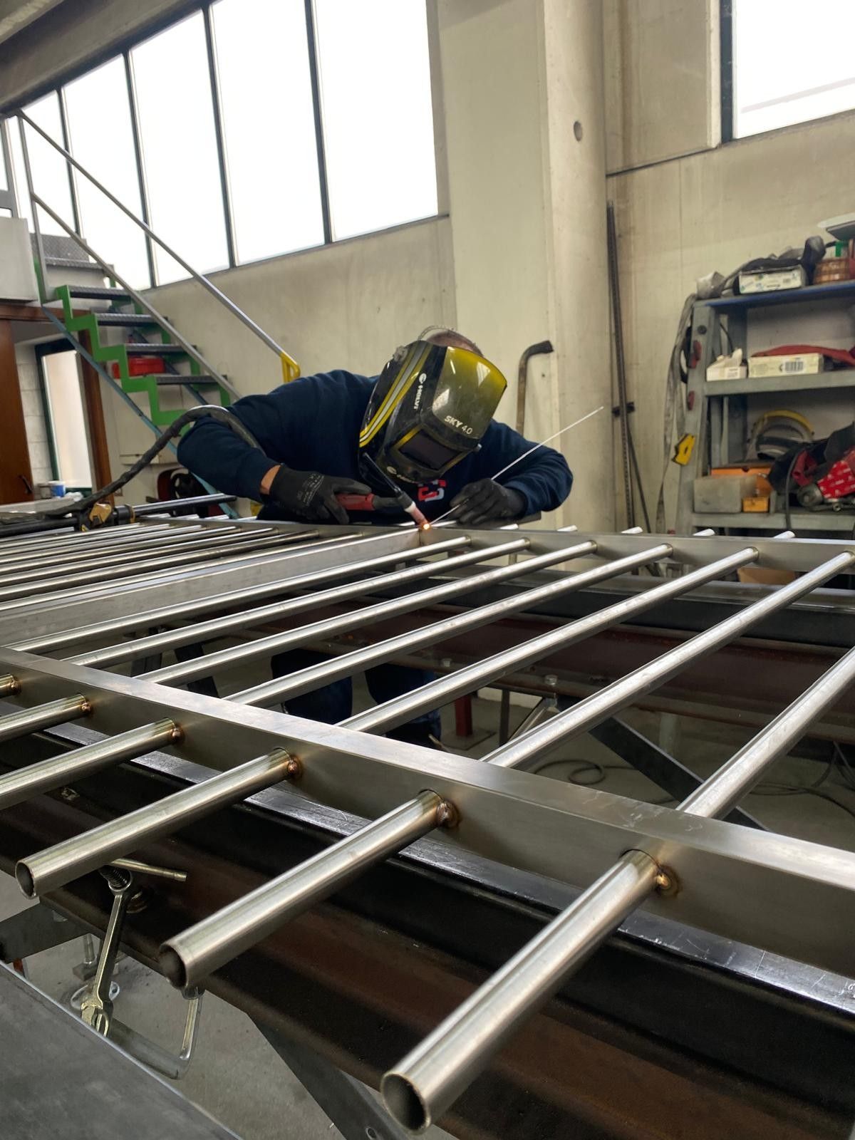 Un operaio con il casco giallo ripara una ringhiera metallica all'interno di un'officina industriale.