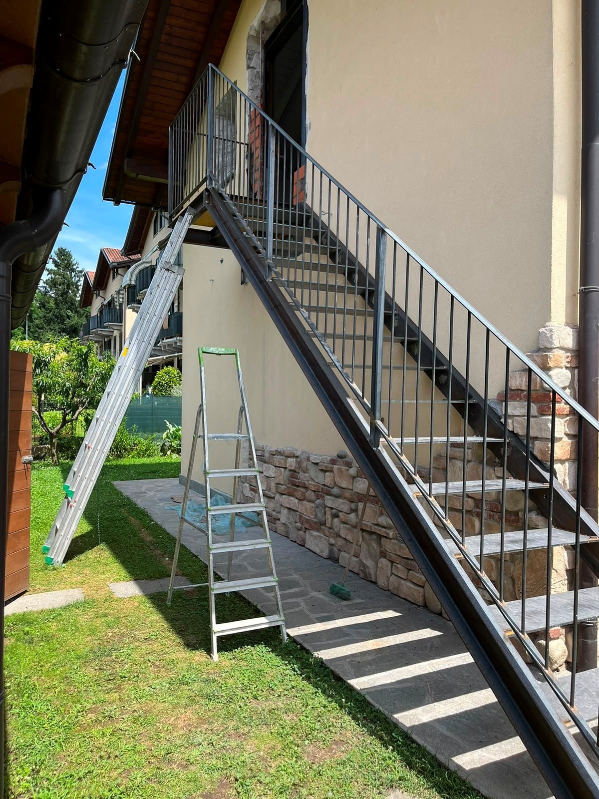 Scala metallica addossata a una porta del secondo piano, accanto a un edificio beige, con una piccola scala a pioli e un cortile erboso sottostante.