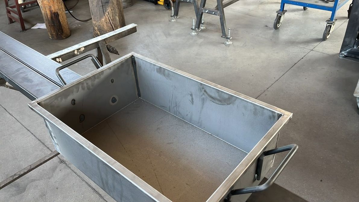 Grande contenitore metallico rettangolare con maniglie laterali, parzialmente riempito d'acqua, in un'officina o in un ambiente di fabbrica.