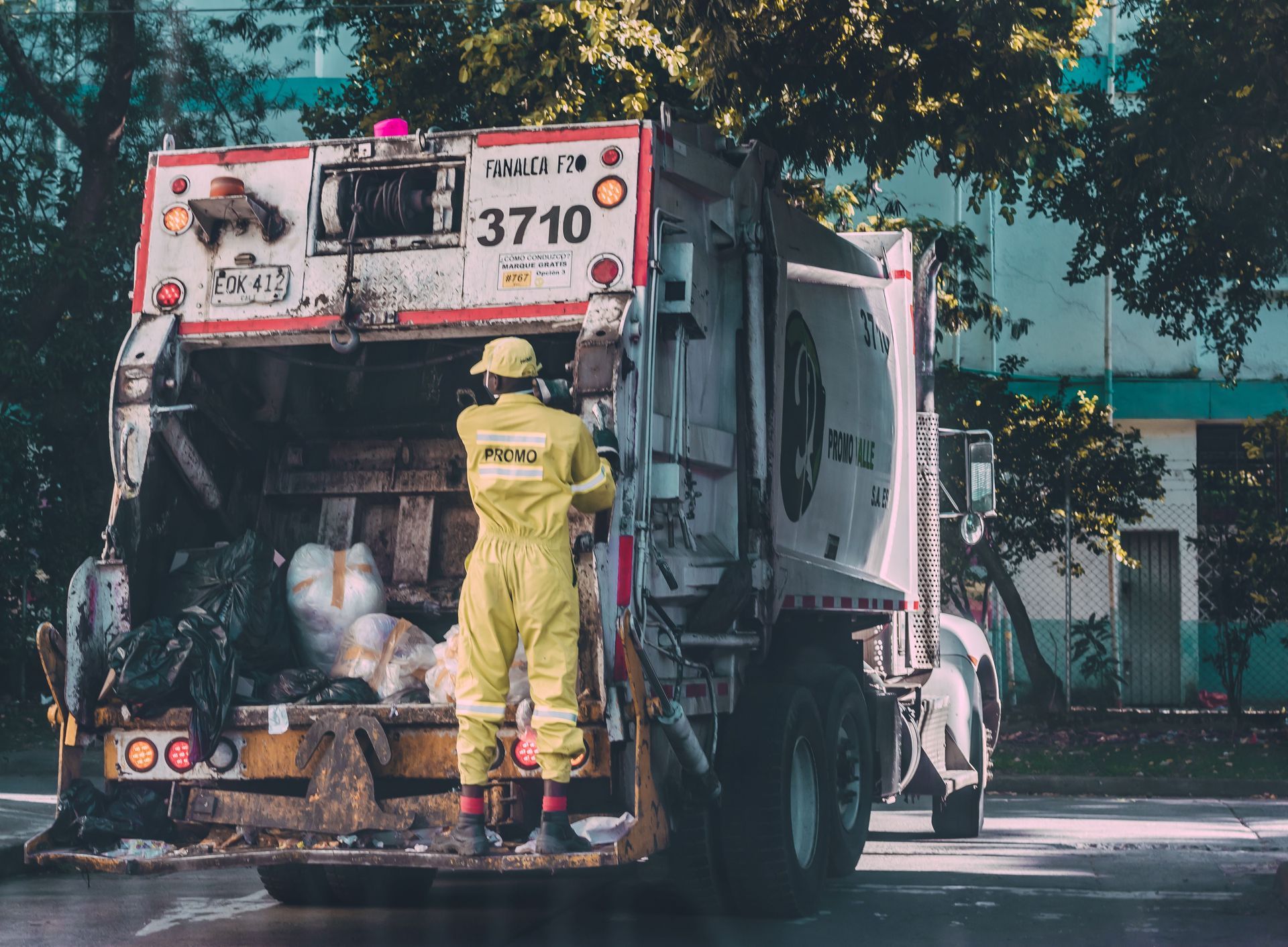 Worker in yellow suit standing on a garbage truck, loading waste bins on a city street.