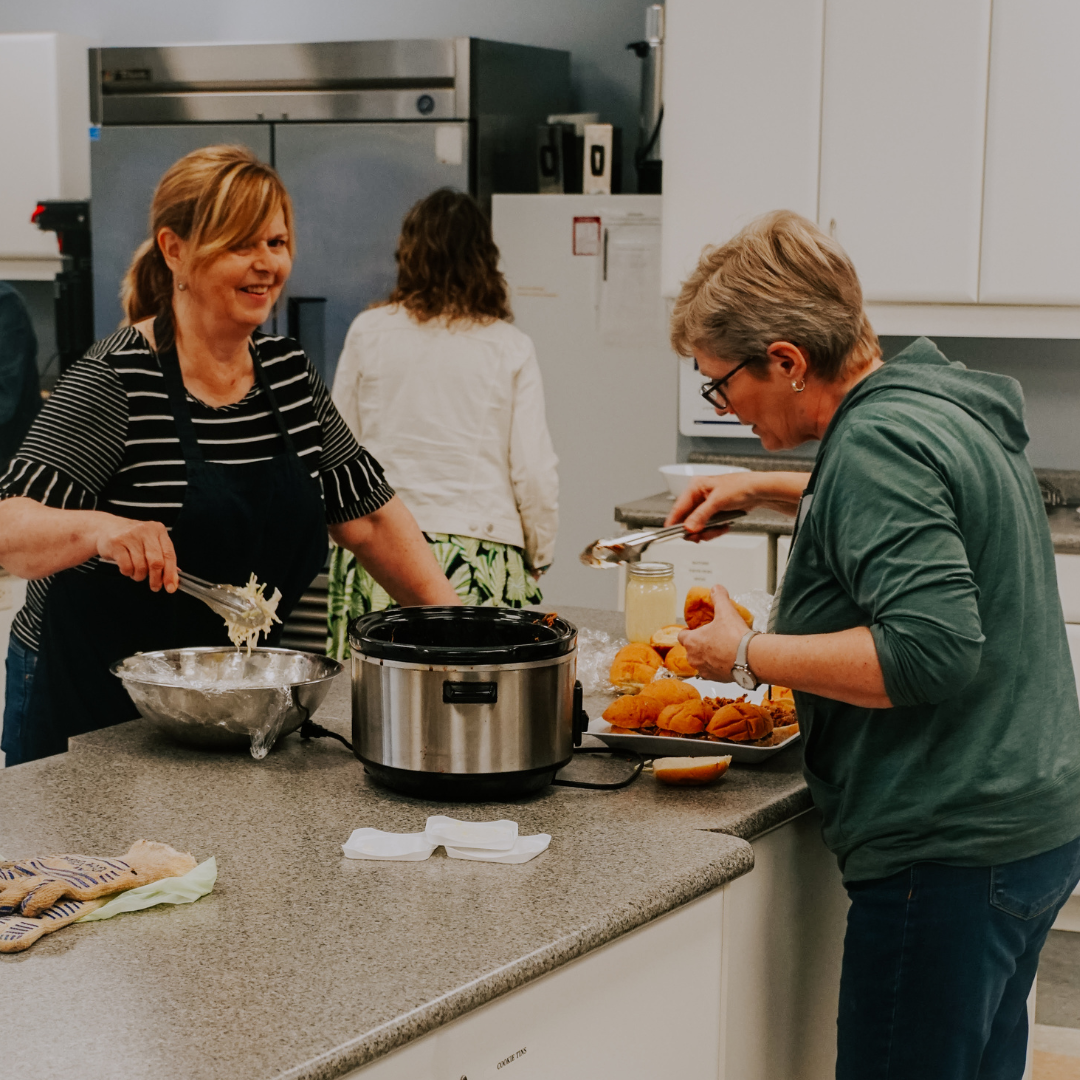 Two women cooking in a kitchen, one stirring, one with food. A slow cooker sits on the counter.